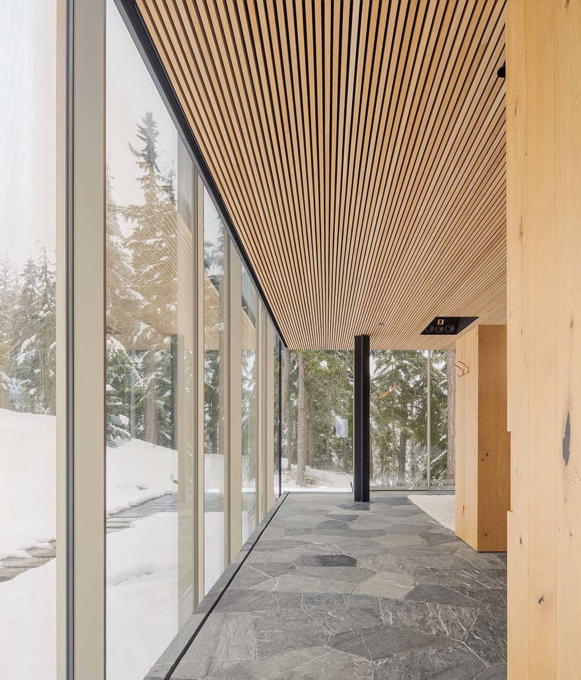 Modern interior corridor with glass walls, wooden ceiling panels, and stone flooring, featuring winter scenery with snow-cove