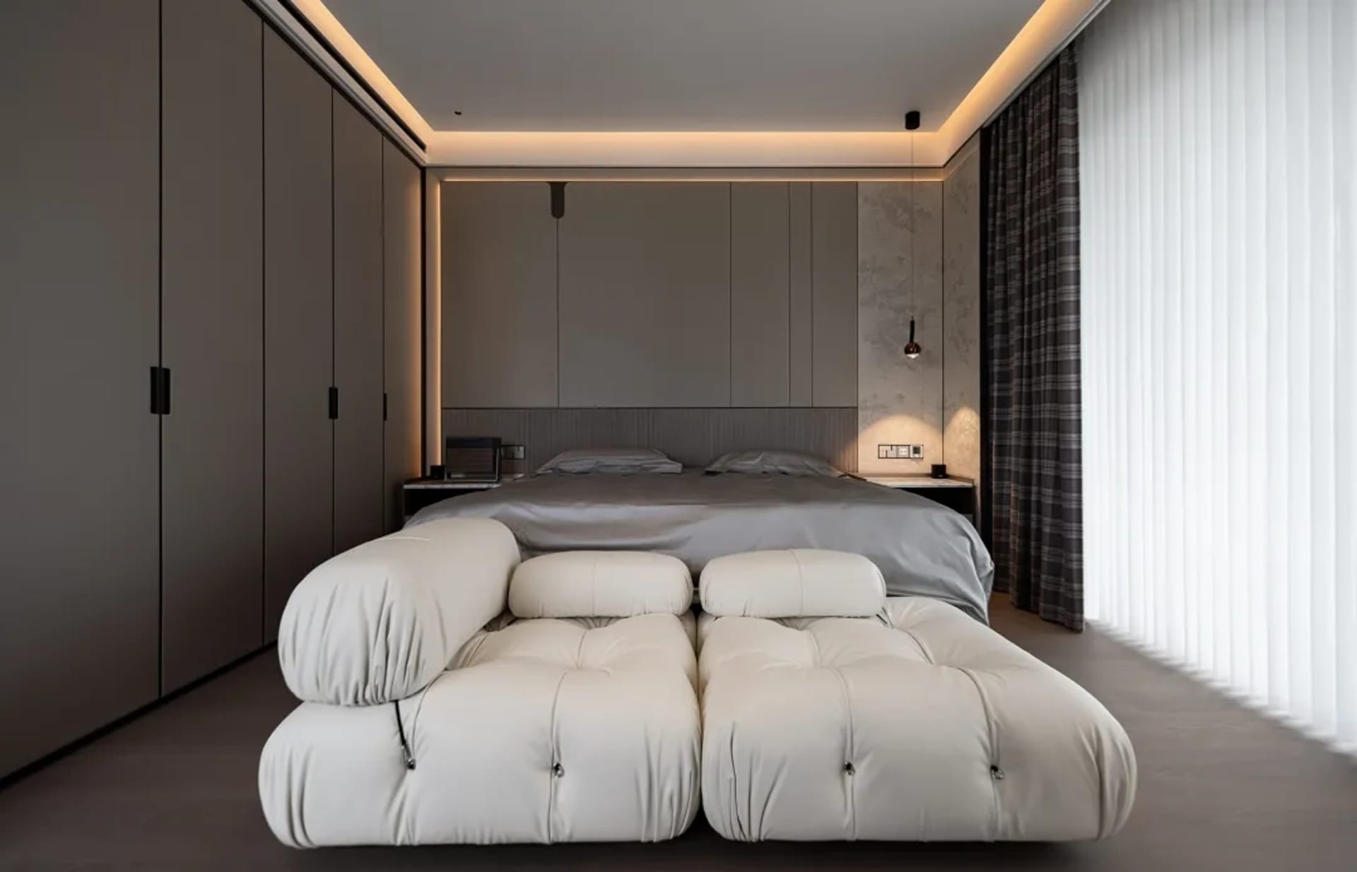 A modern minimalist bedroom featuring a large bed with a tufted headboard, a white fabric bench, a large window with vertical