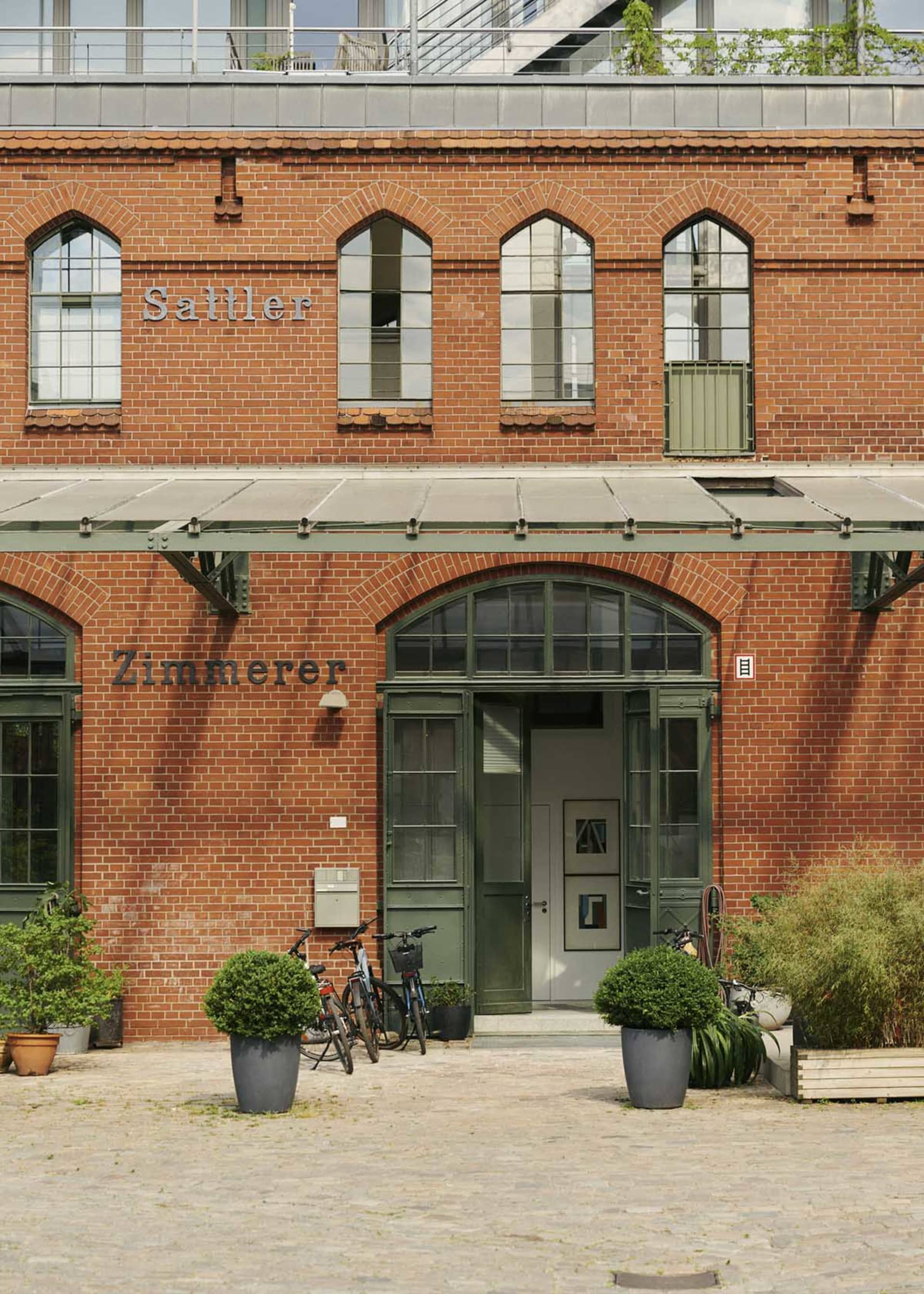 A rustic, industrial building with red brick walls and green window frames, featuring the words 'Sattler Zimmerer' above the
