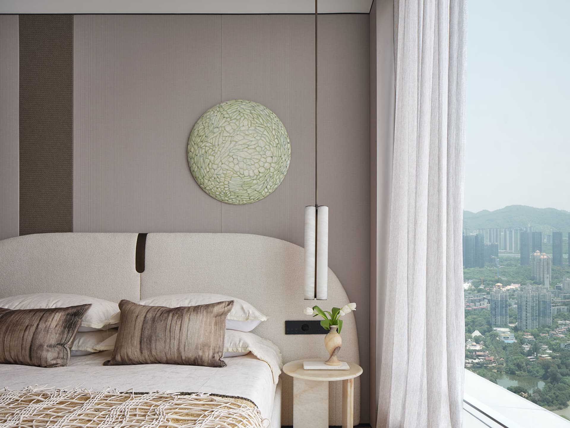 Modern bedroom featuring textured fabric bed, side table with vase and book, and round decorative artwork on the wall.
