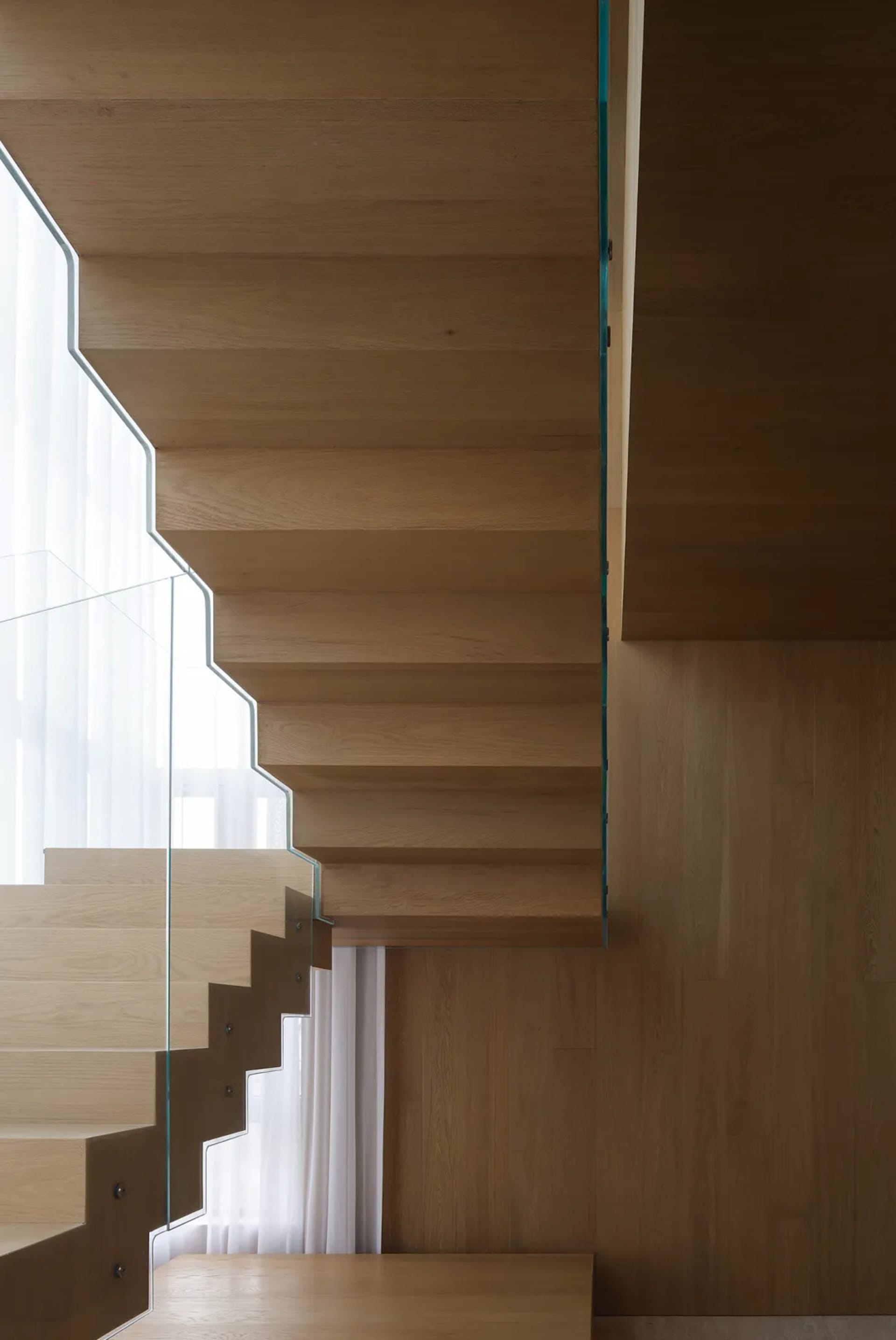 Modern wooden staircase with glass railings and white curtains in the background.