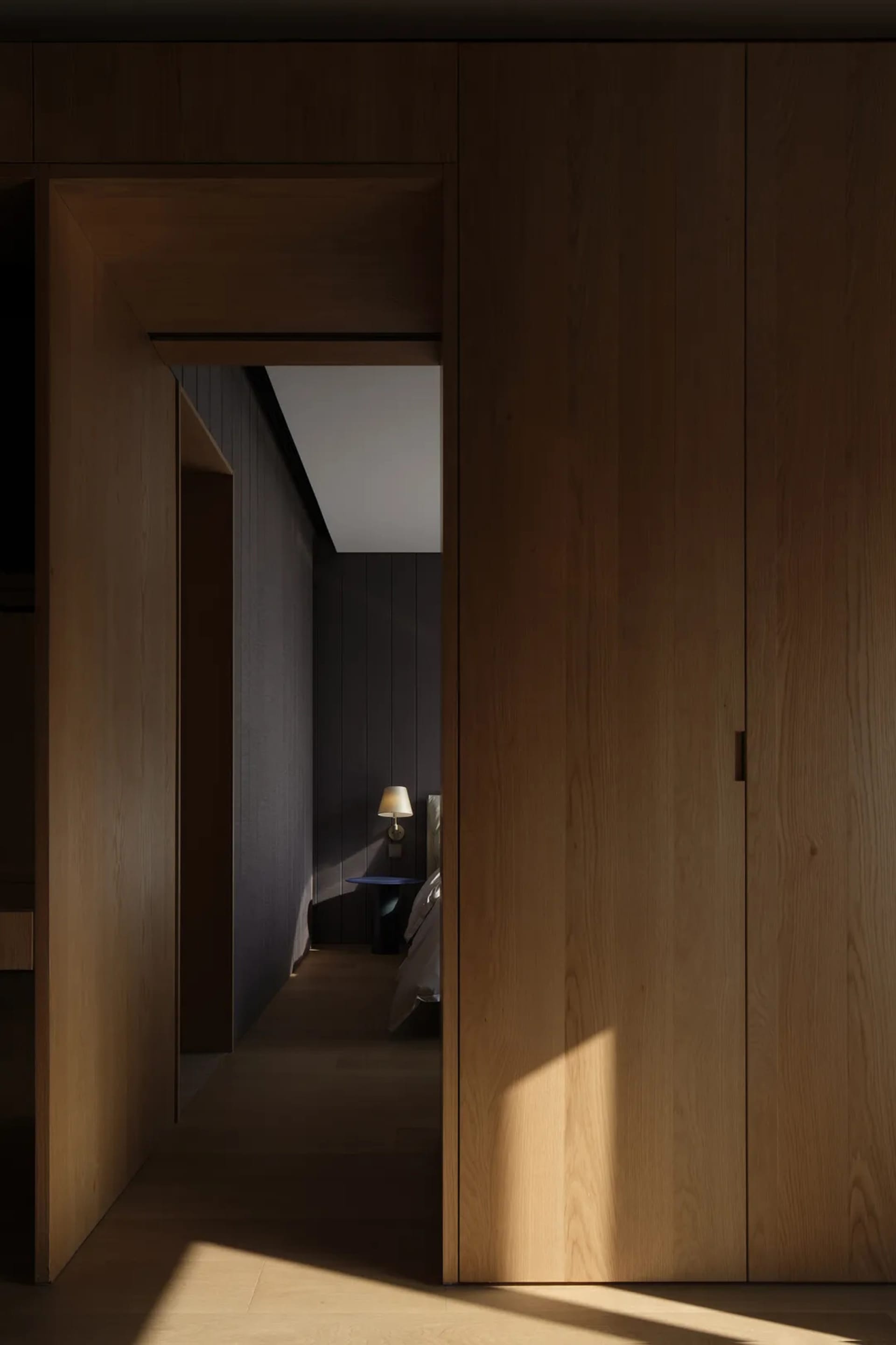 A modern, minimalist wooden doorway with a lamp in the adjacent bedroom.