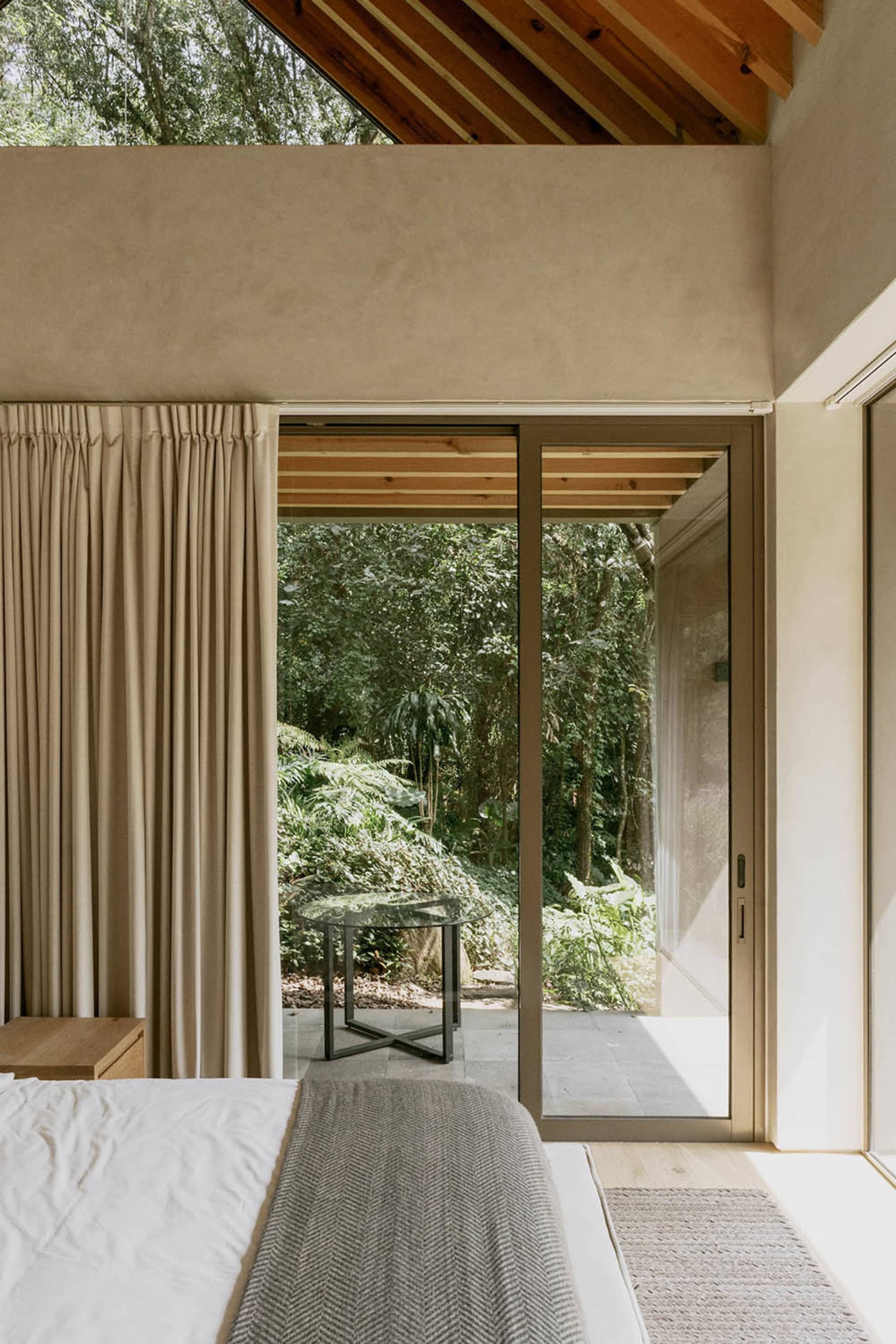 A modern bedroom featuring concrete, glass, and wooden materials, with a view of the forest through large windows.