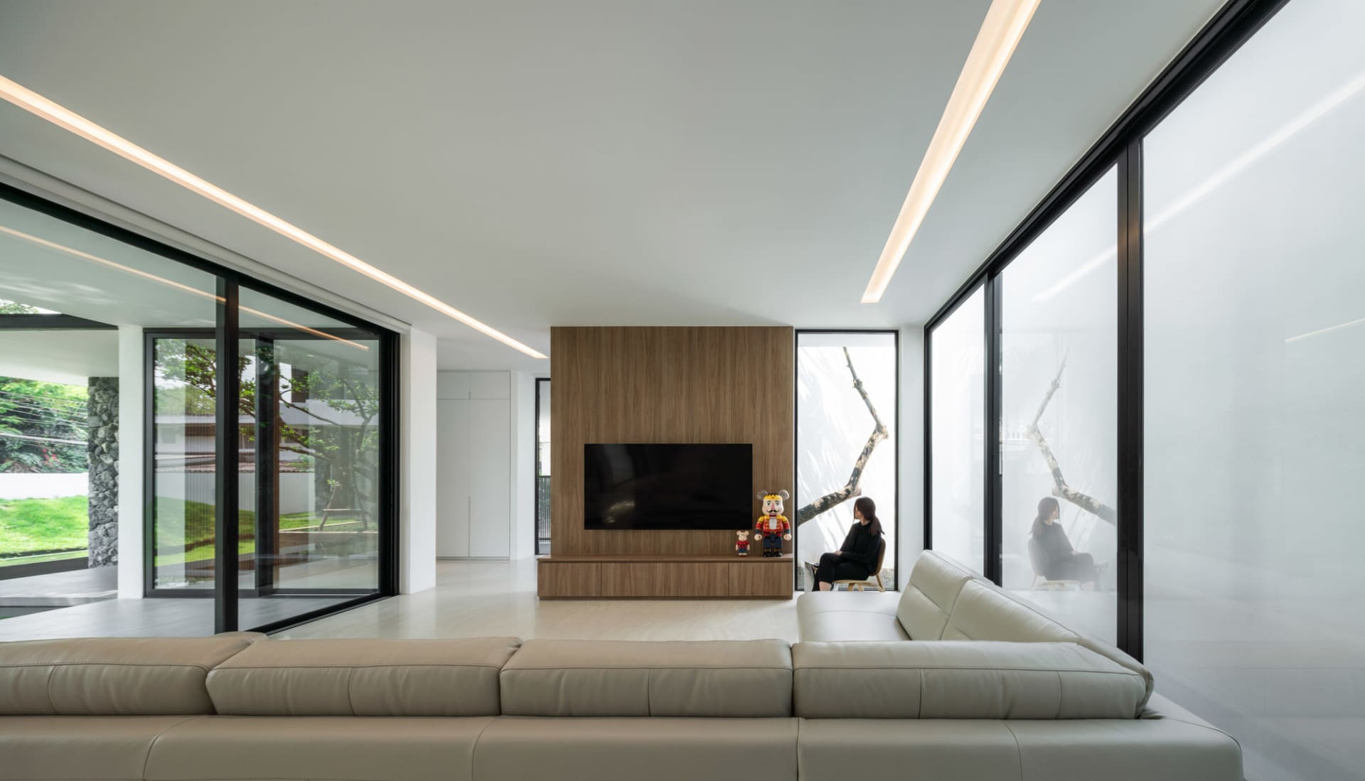 A modern living room with a large beige leather sofa, a wood-paneled wall with a television, and glass windows.