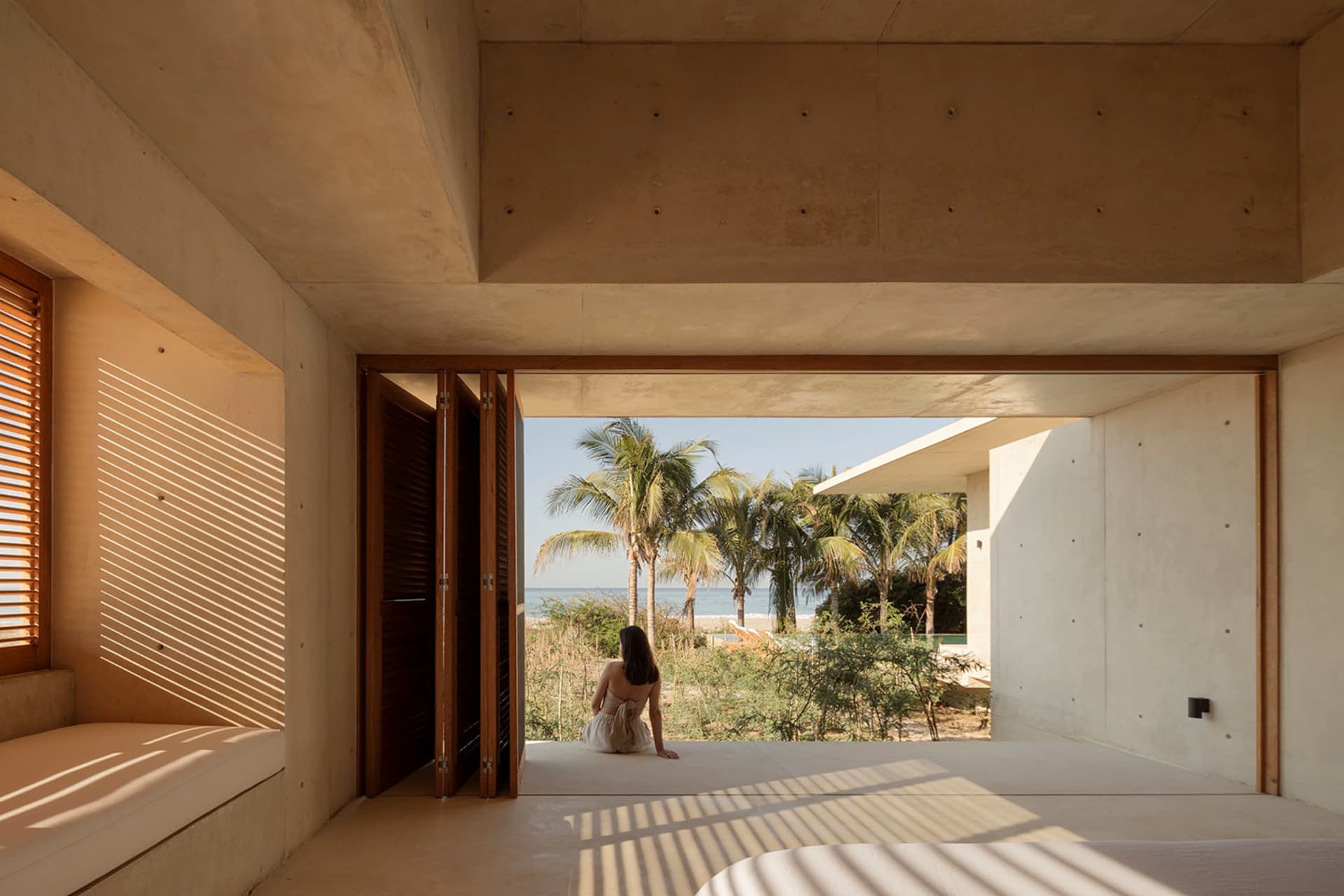 A modern residential bedroom with concrete walls and floor, featuring a view of the beach through an open window.