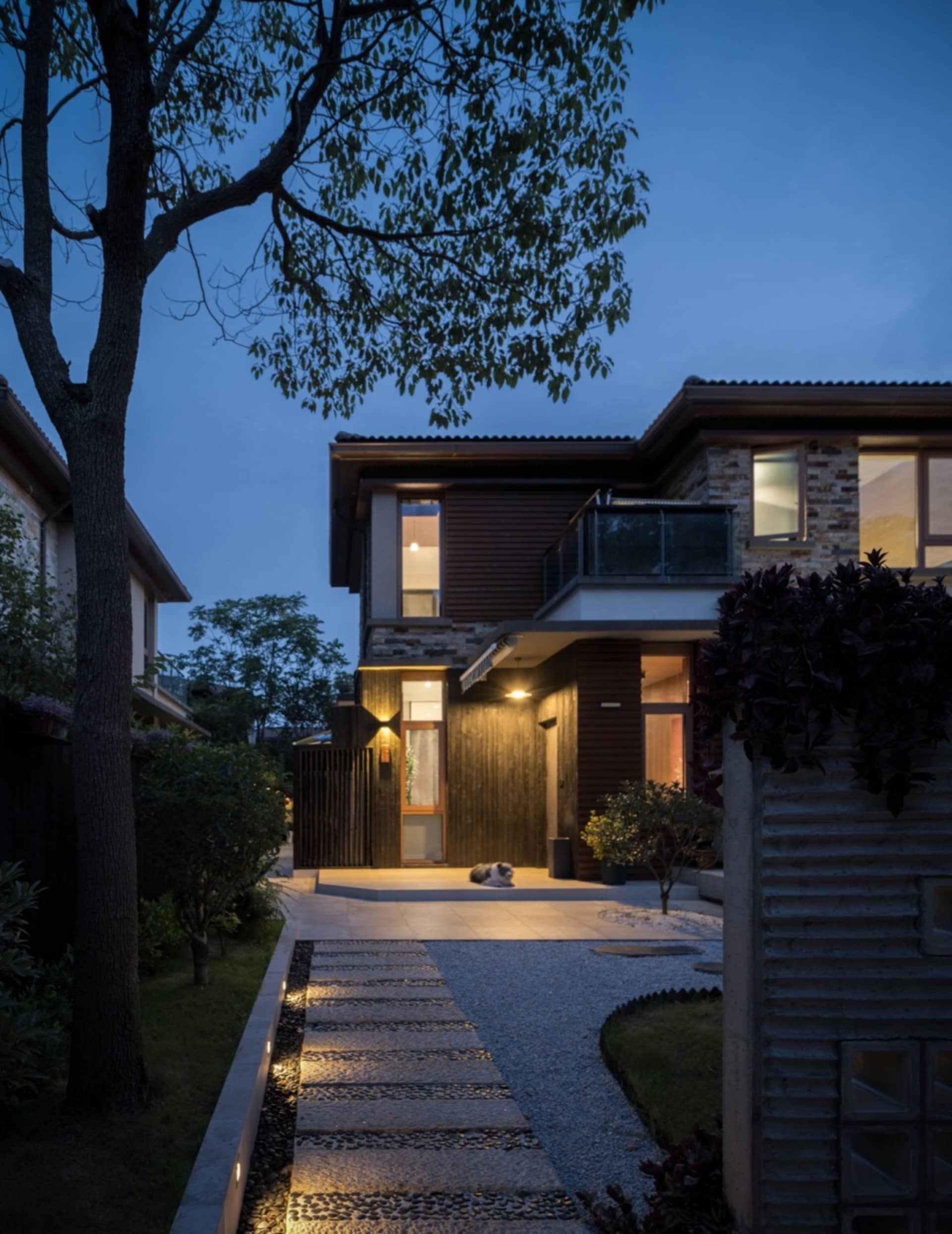 A modern residential home with wooden elements and brick walls at dusk, featuring a lit entranceway and a path leading to the