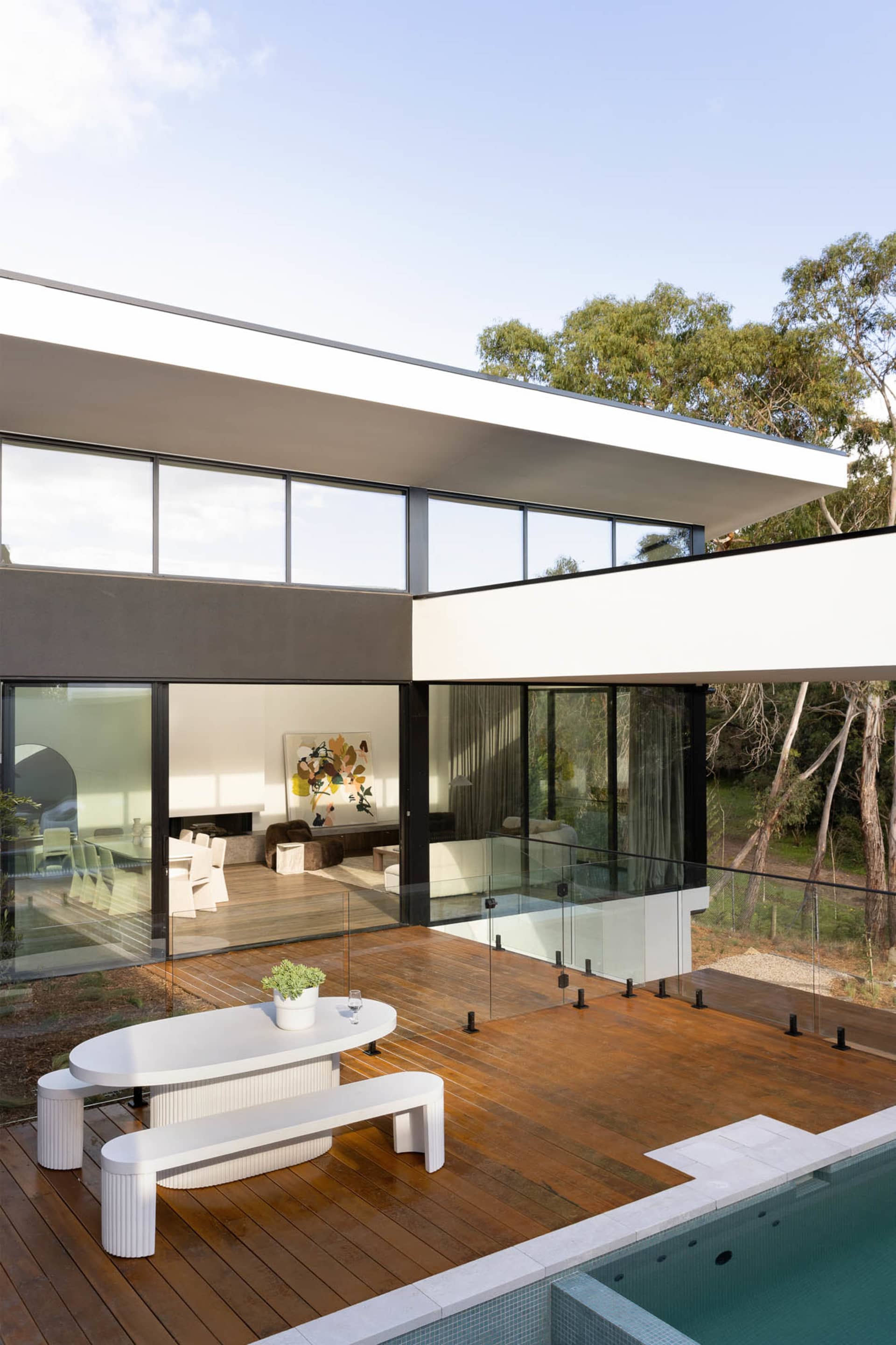 Modern residential exterior featuring wooden deck with white bench and table, glass railing, and swimming pool.