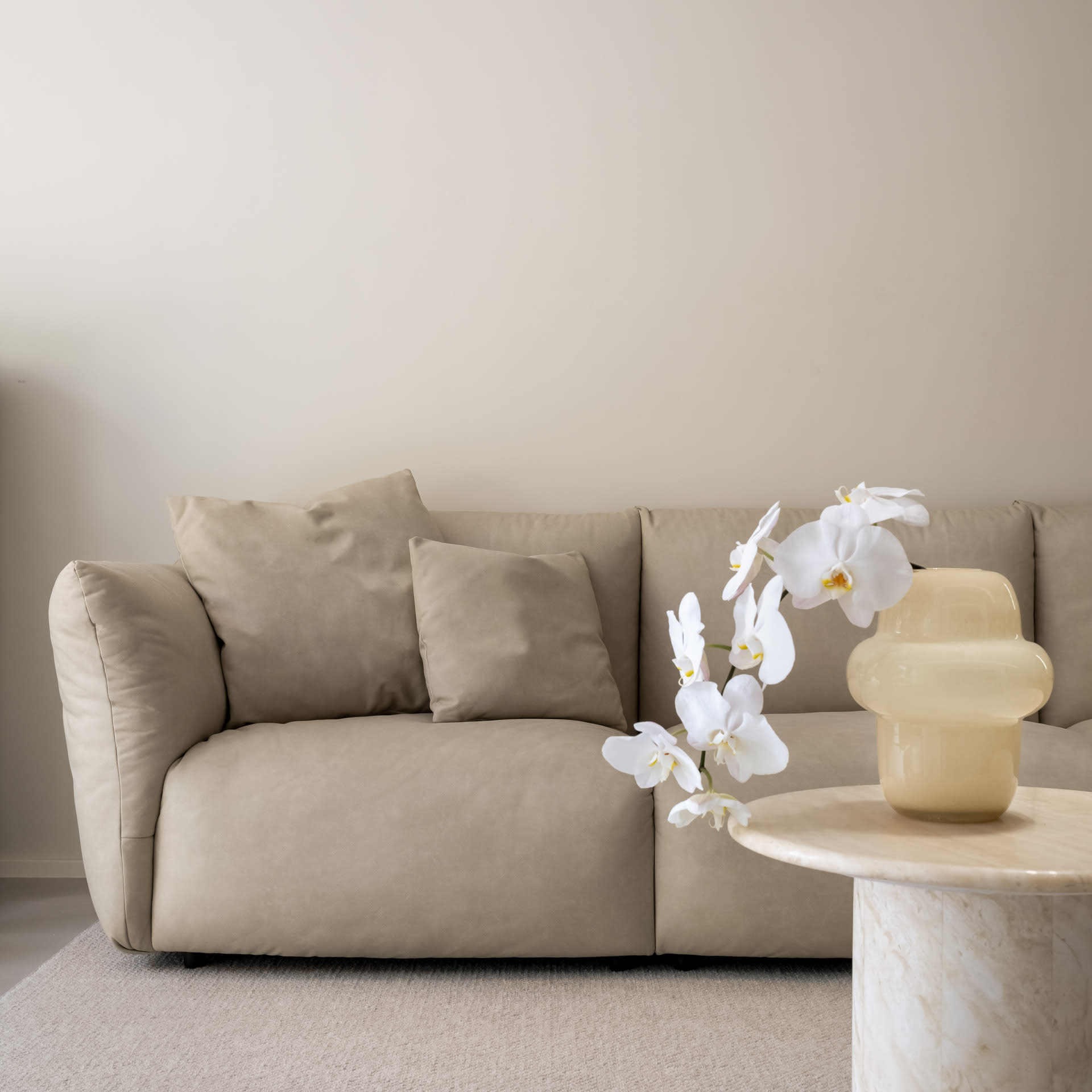 A comfortable, minimalist living room interior with a beige leather sofa, white orchid flowers in a vase, and a round marble