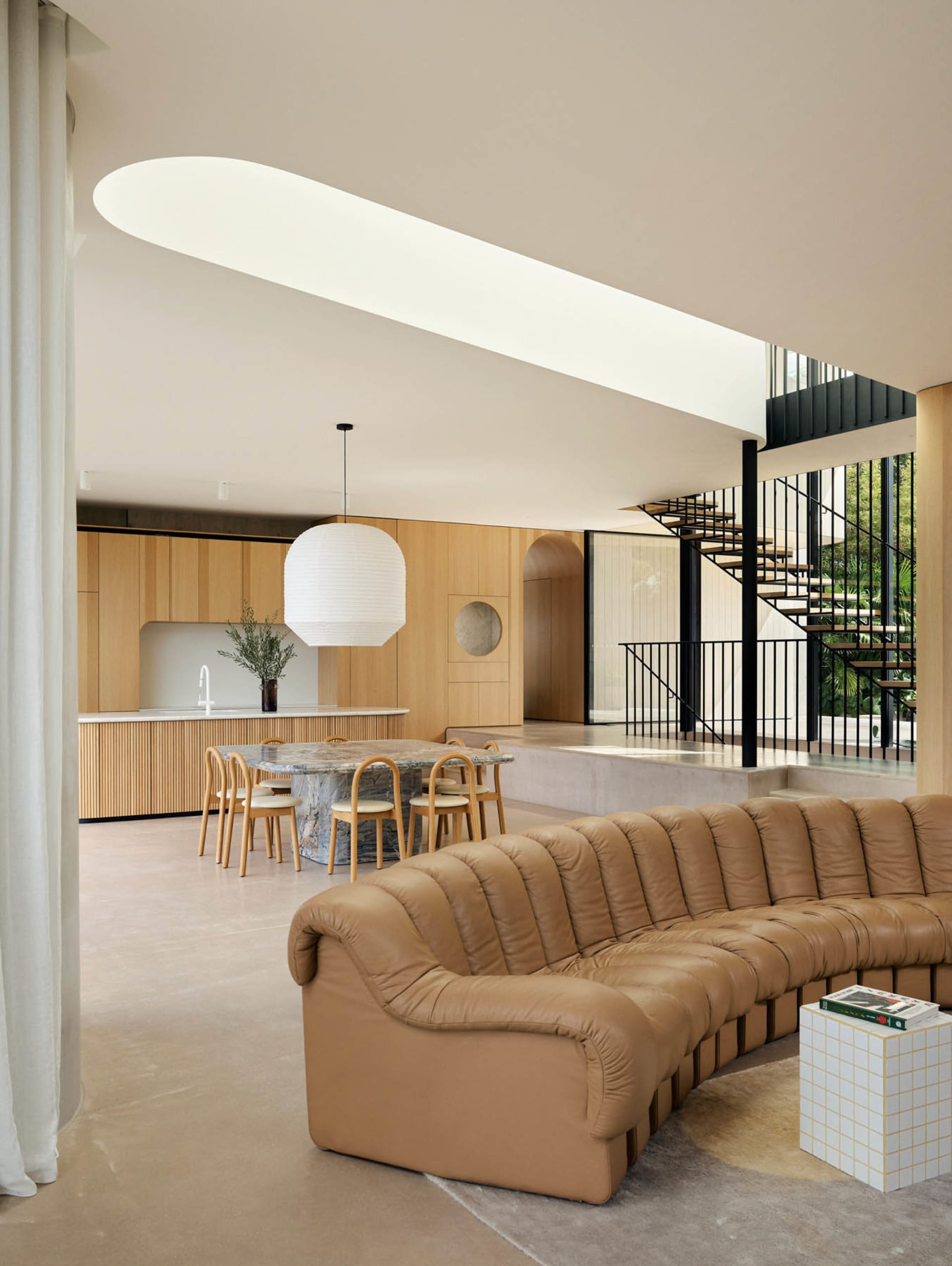 A modern living room features a brown leather sofa, a large white pendant light, and a kitchen area with wooden cabinets and