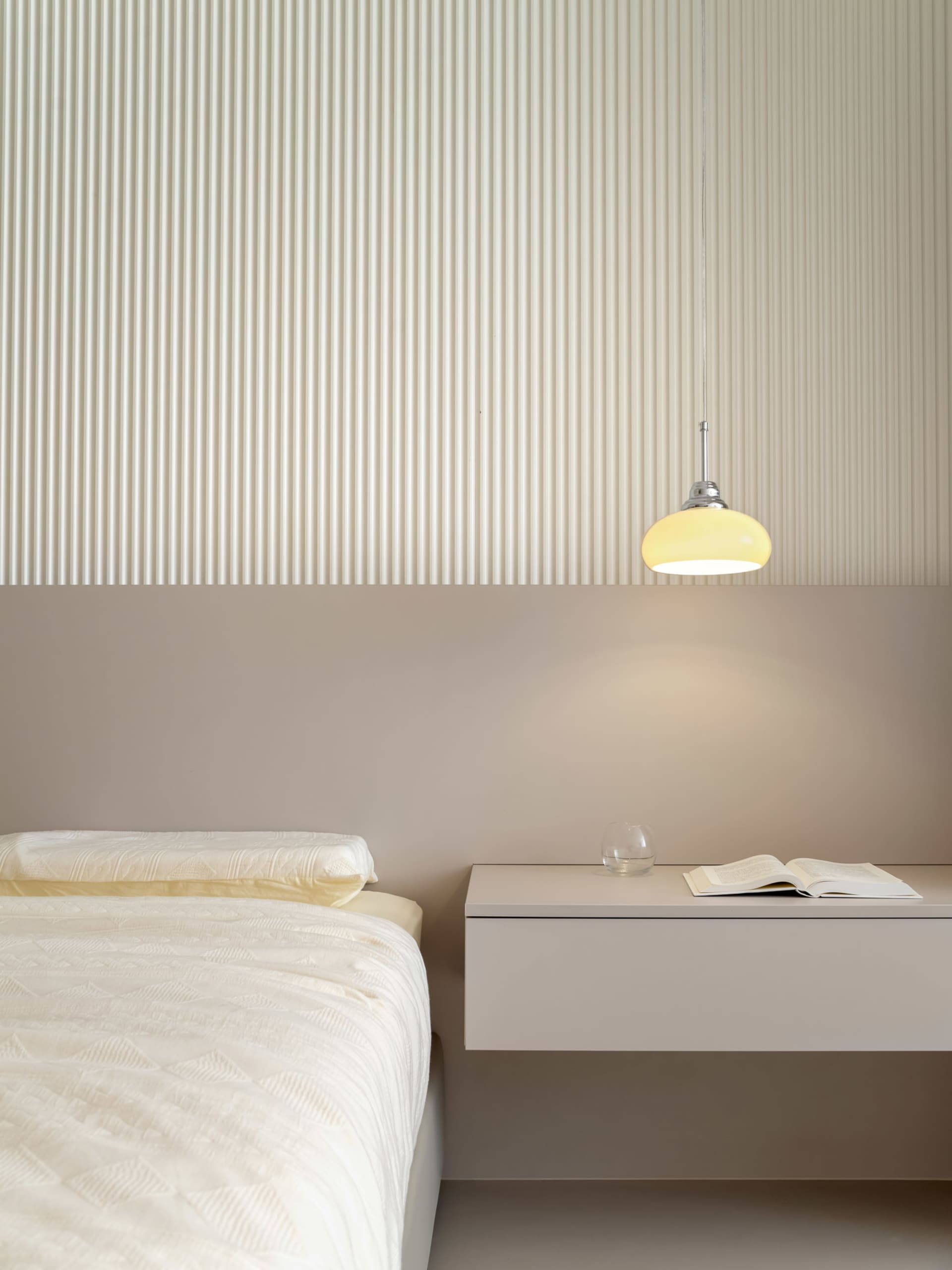 A modern minimalist bedroom featuring white bedding, a floating nightstand with a glass of water and an open book, and a pend