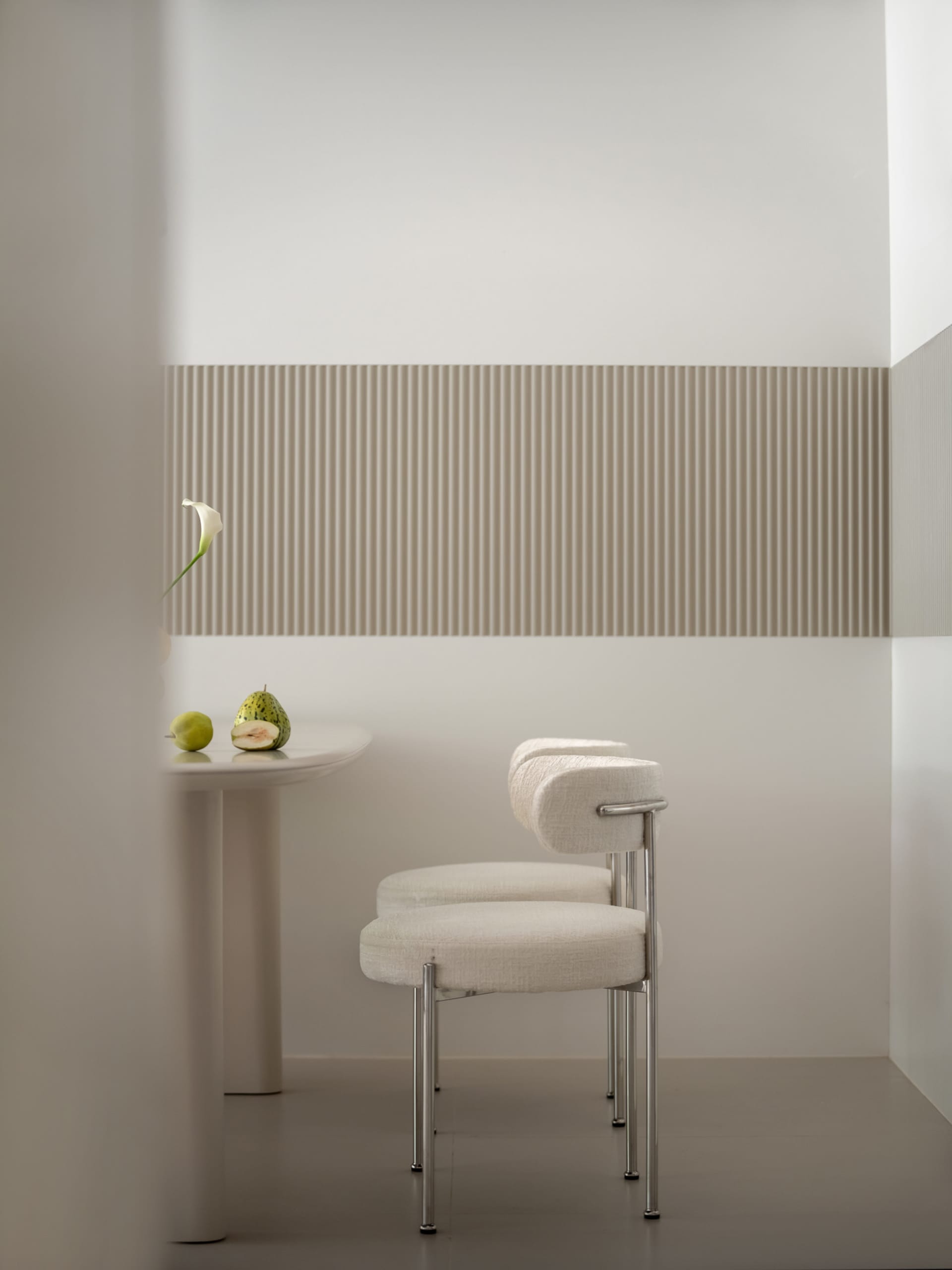 A modern white interior featuring fabric chairs, a table with fruits, and a striped wall.