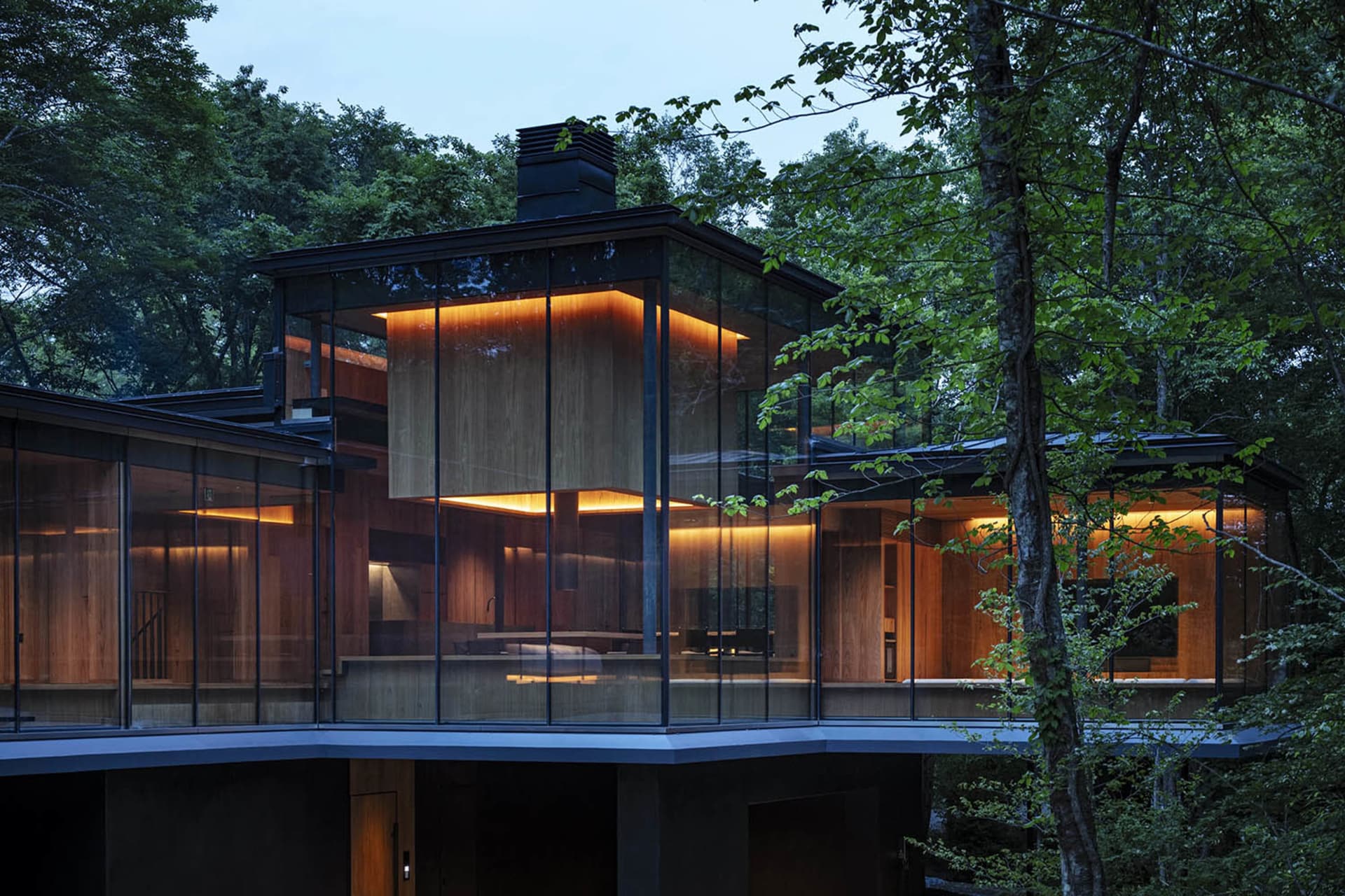 A modern glass house surrounded by trees with warm indoor lights visible through the glass walls.
