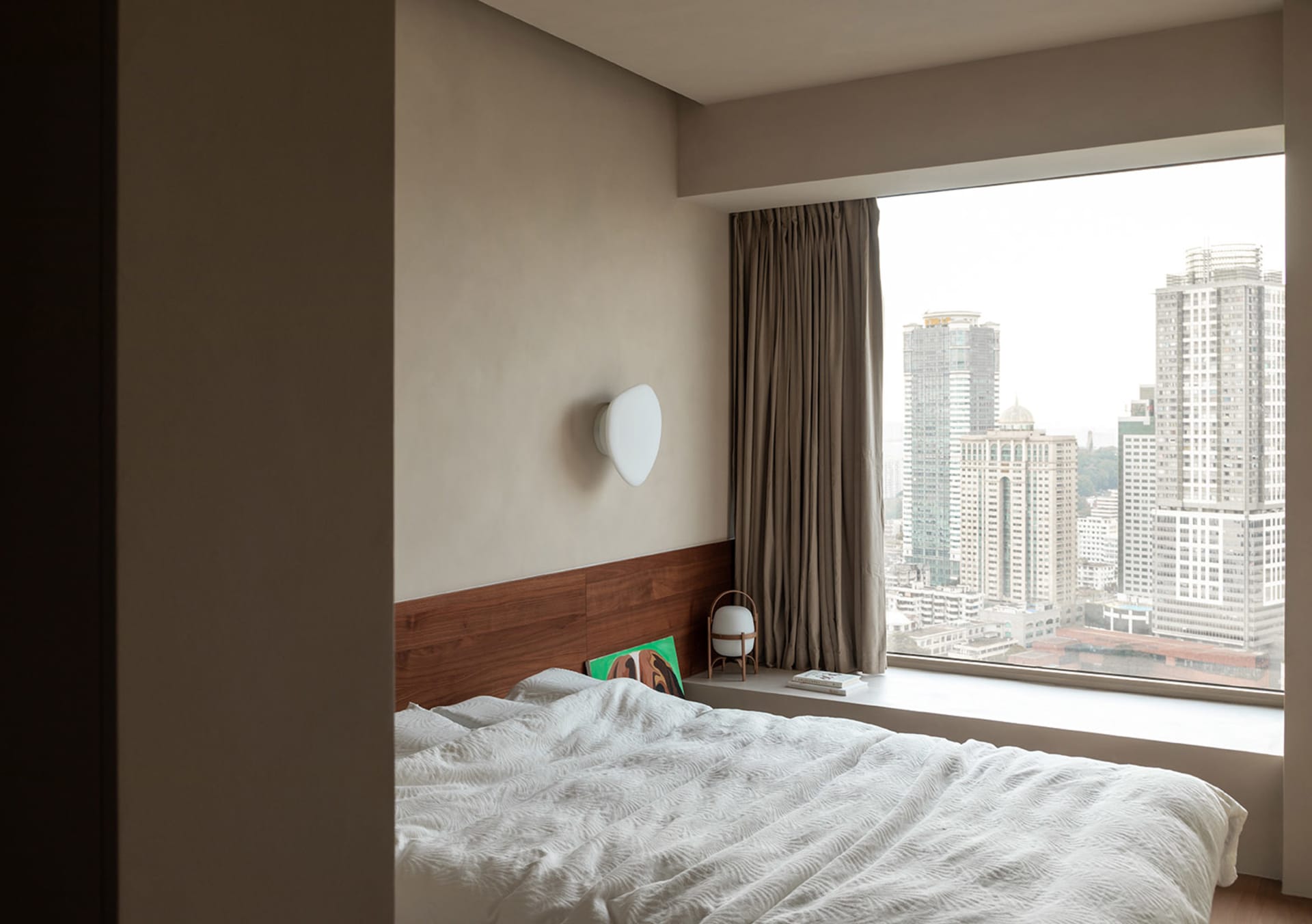 A minimalist bedroom with a large window overlooking the city, featuring a blank white blanket on the bed and a wall sconce.