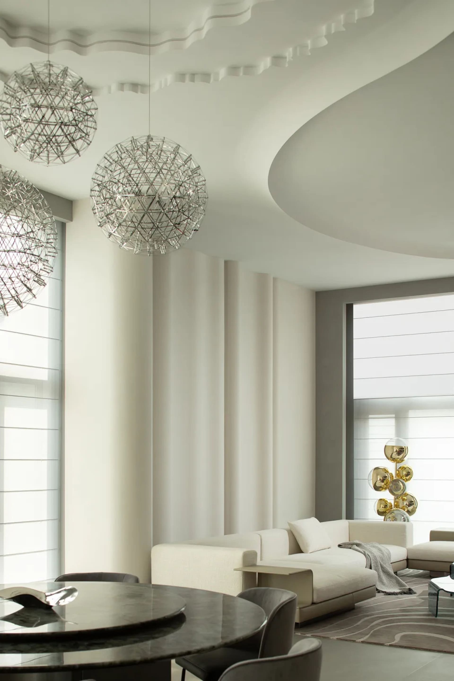 A modern minimalist living room with white sofas, floor-to-ceiling windows, and metallic pendant lights hanging from the ceil