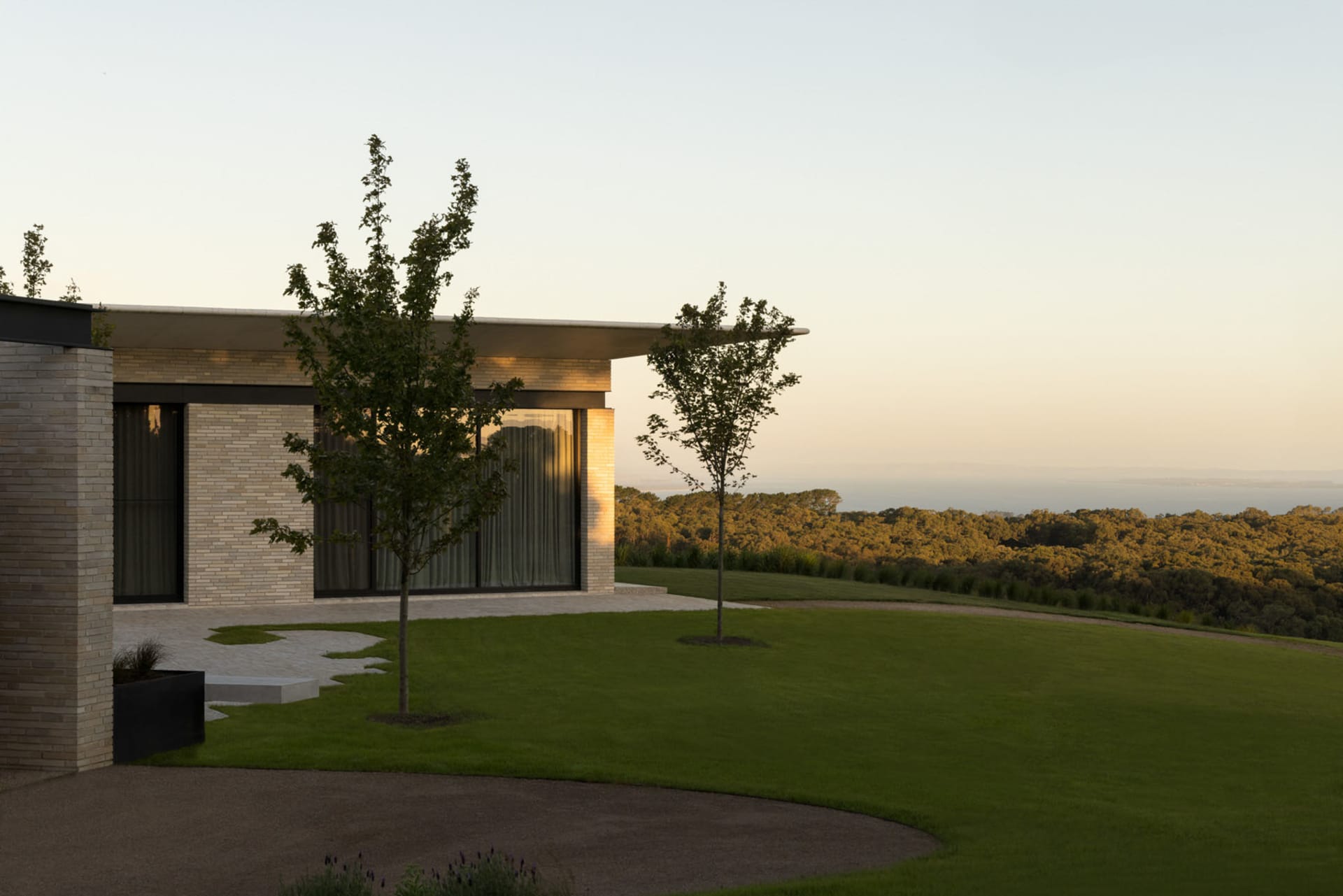 Modern residential exterior with brick walls, glass windows, and a landscaped yard overlooking a forested area during sunset.