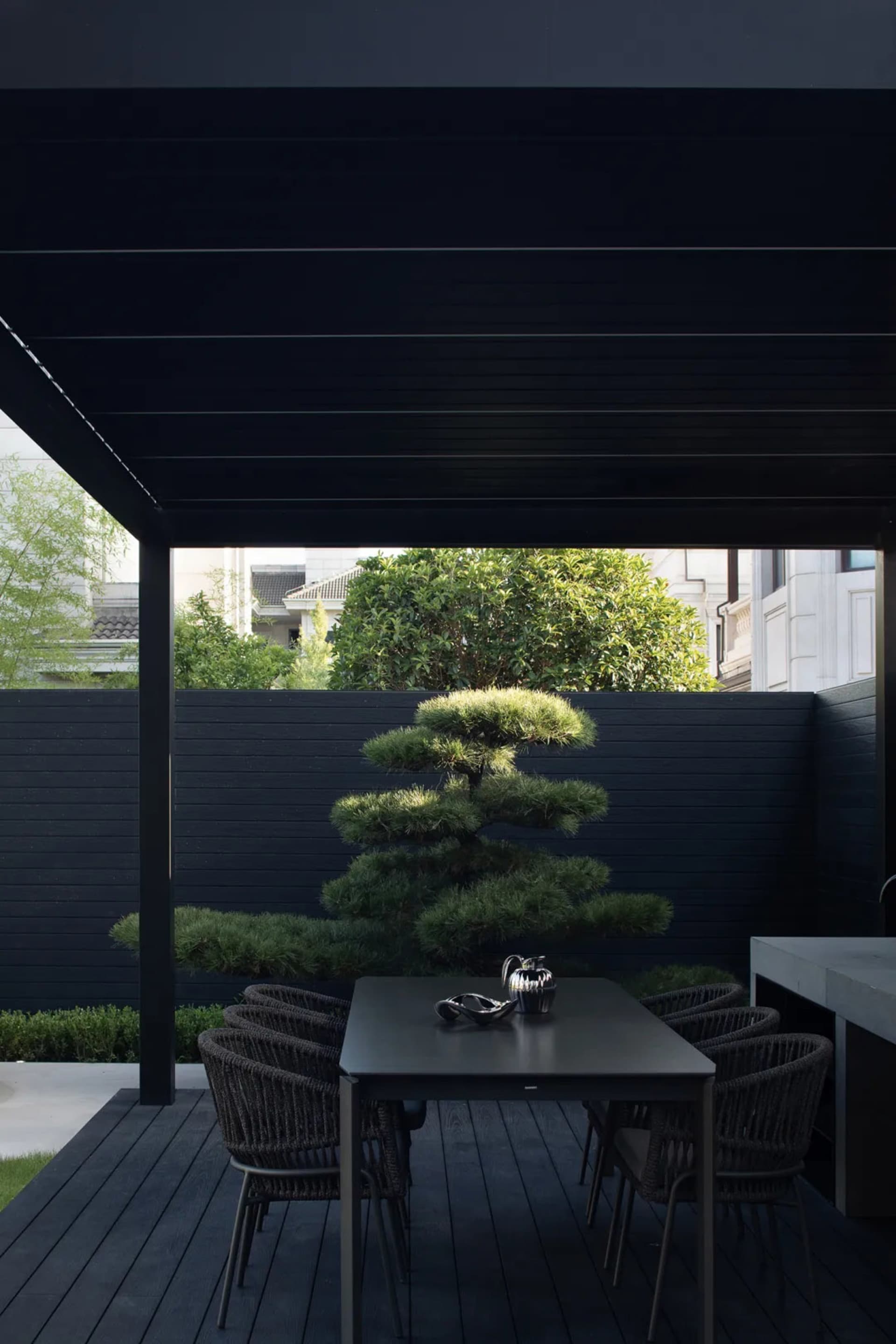 A modern outdoor dining area featuring a dark wooden deck with a black pergola and a black table surrounded by black chairs,
