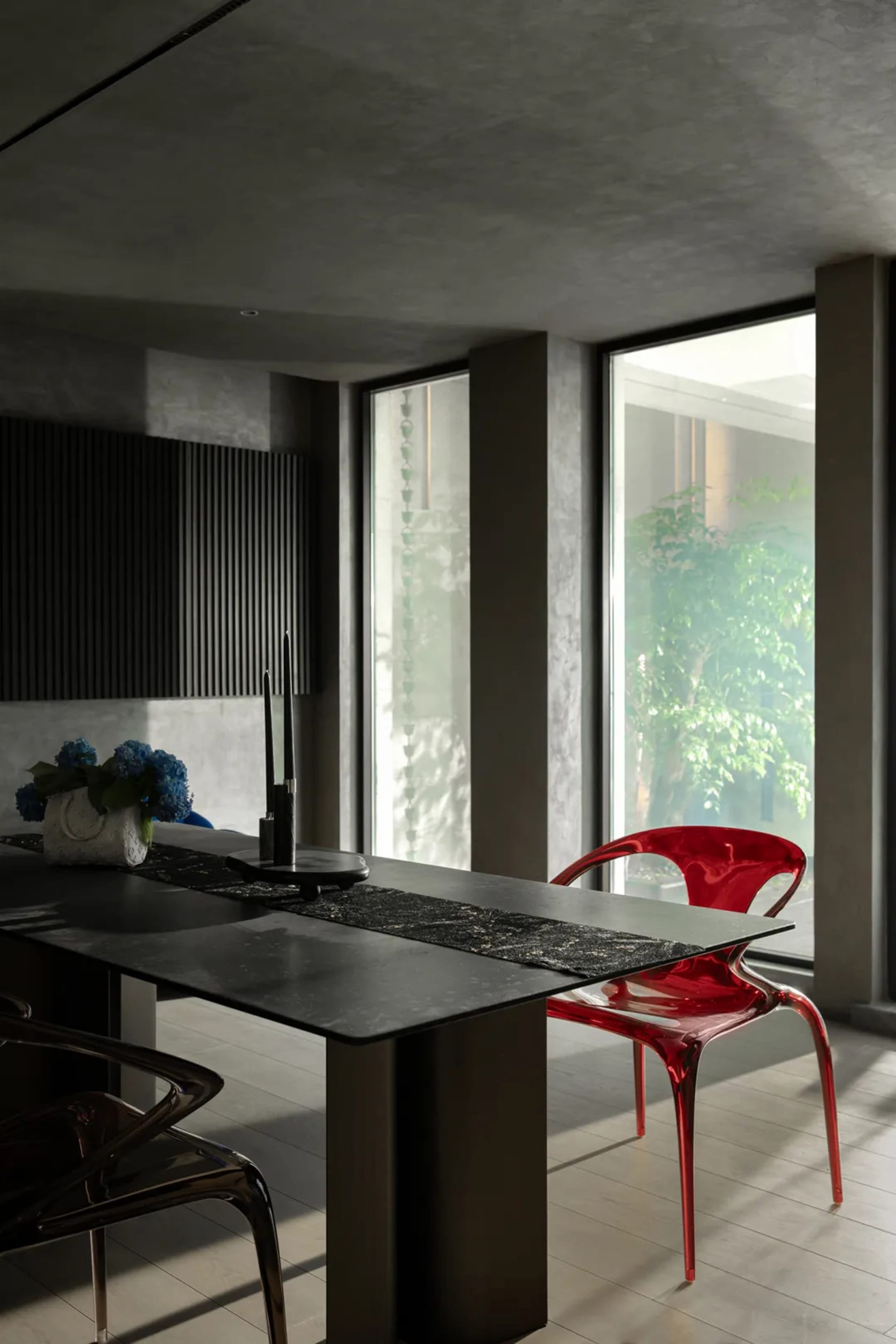 A modern dining room with a dark metal dining table and a red acrylic chair, featuring a black table runner and blue flowers.