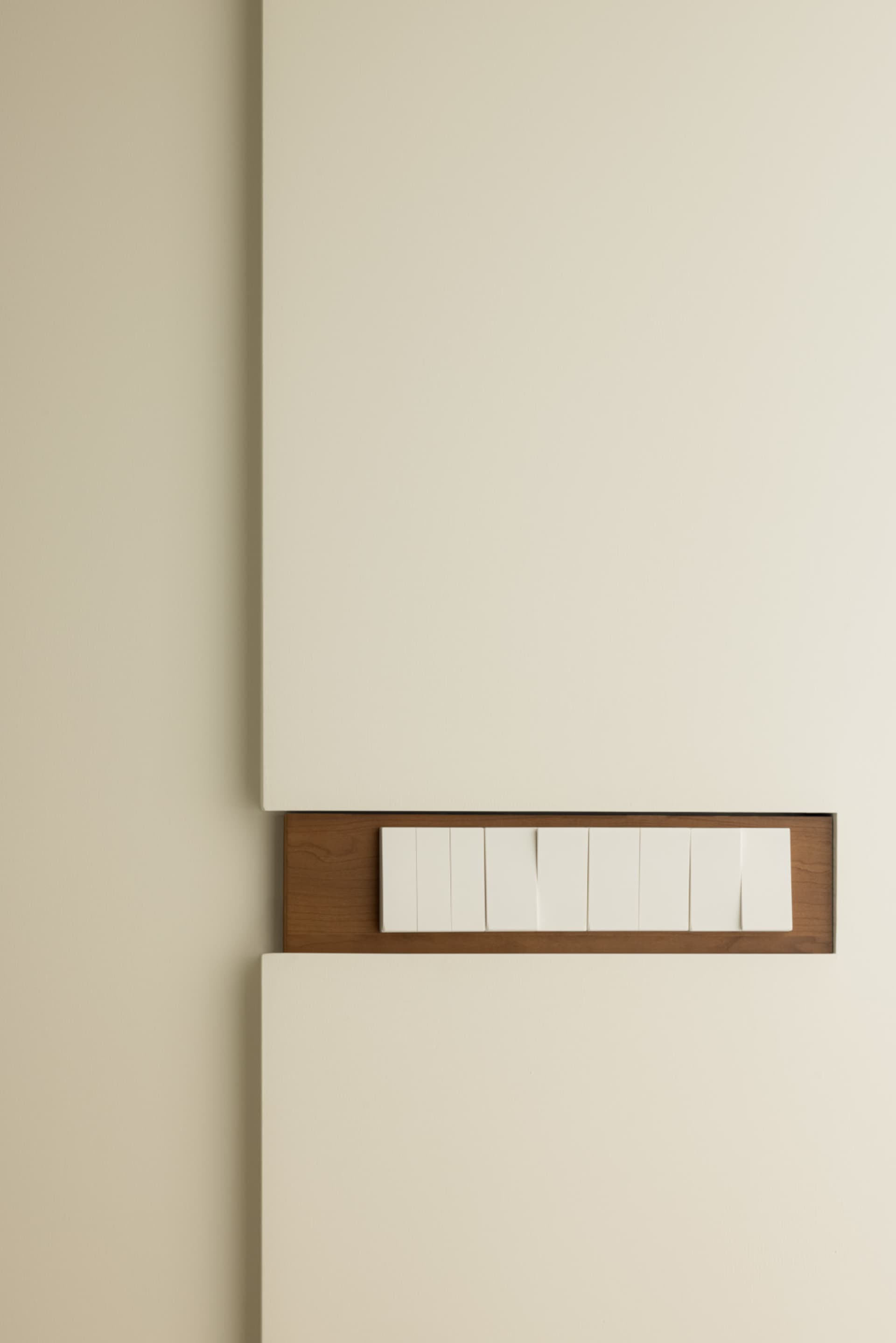 A minimalist light switch panel with a wooden frame and white plastic buttons mounted on a beige wall.