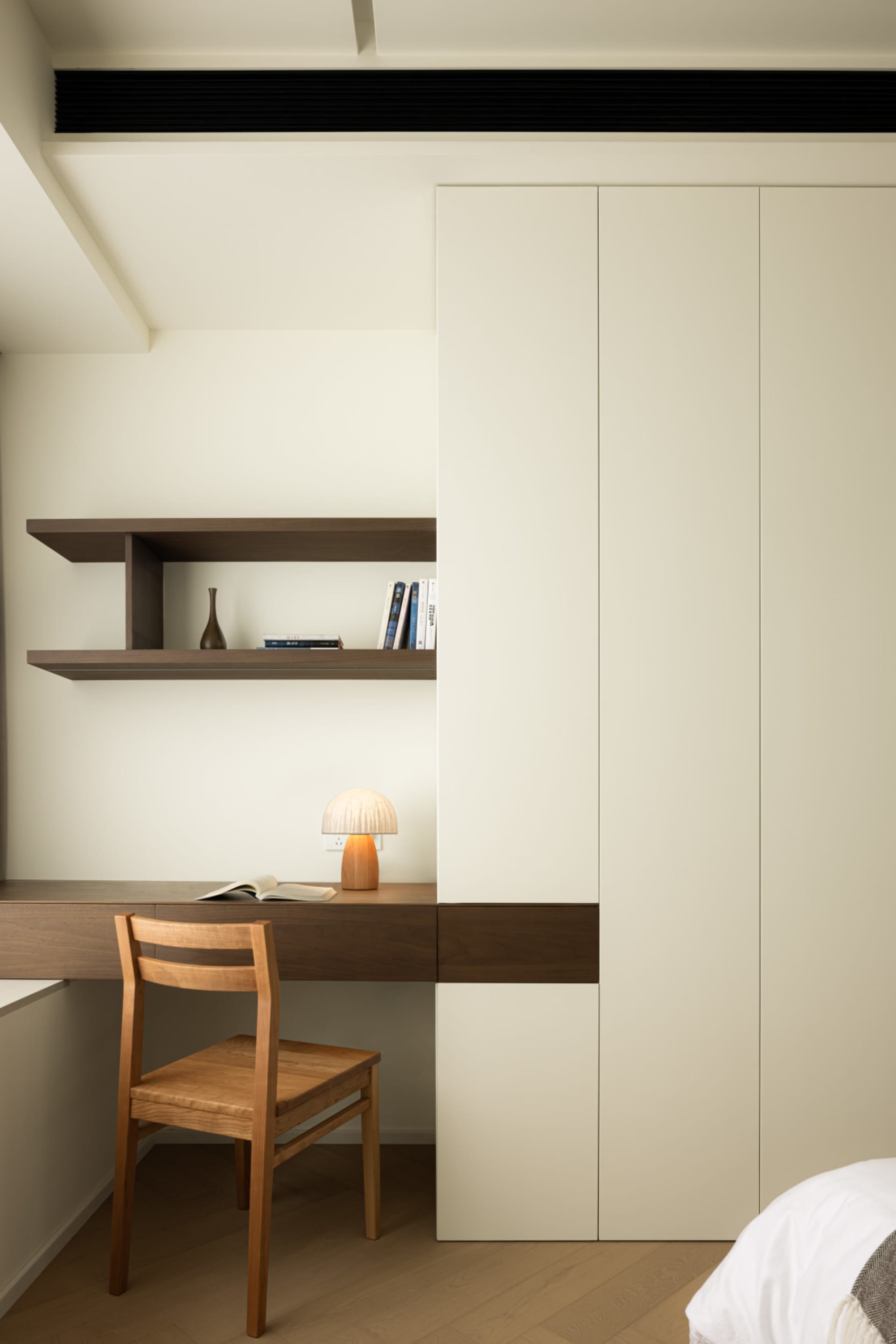 A minimalist bedroom with a wooden chair, white desk lamp, and white bookshelf.