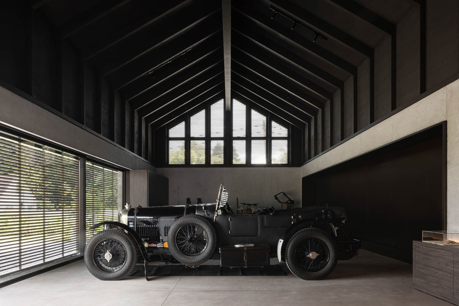 A vintage black car sits in a high-ceilinged, industrial-style garage with concrete walls and floor.
