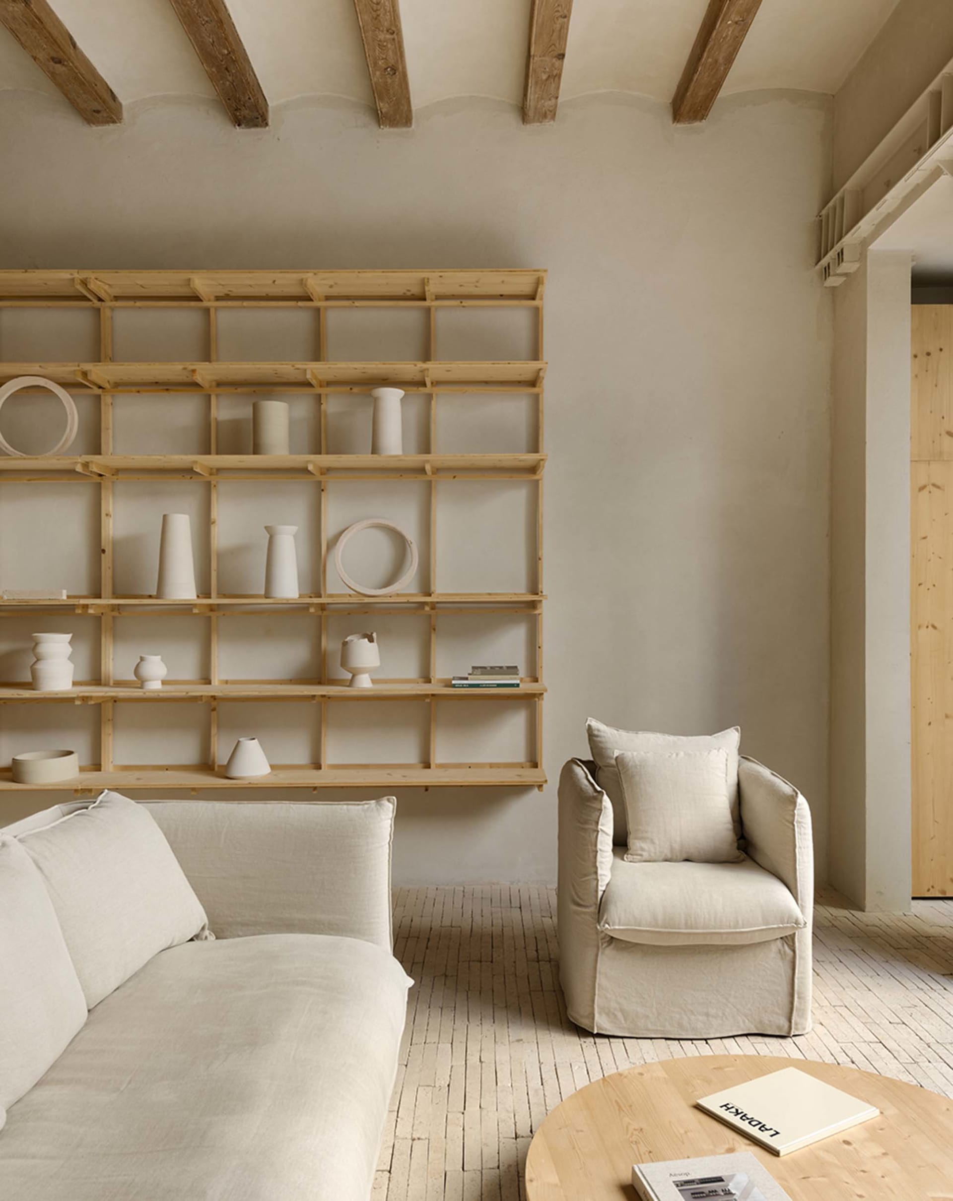Modern minimalist living room with wood shelves, concrete surfaces, and a linen sofa and armchair.