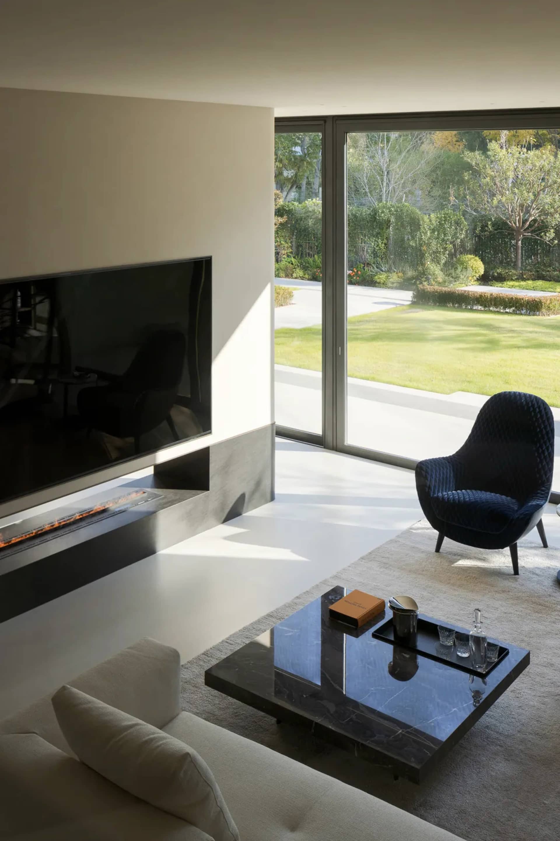 A modern minimalist living room with marble coffee table, concrete fireplace, and glass sliding door leading to a garden.