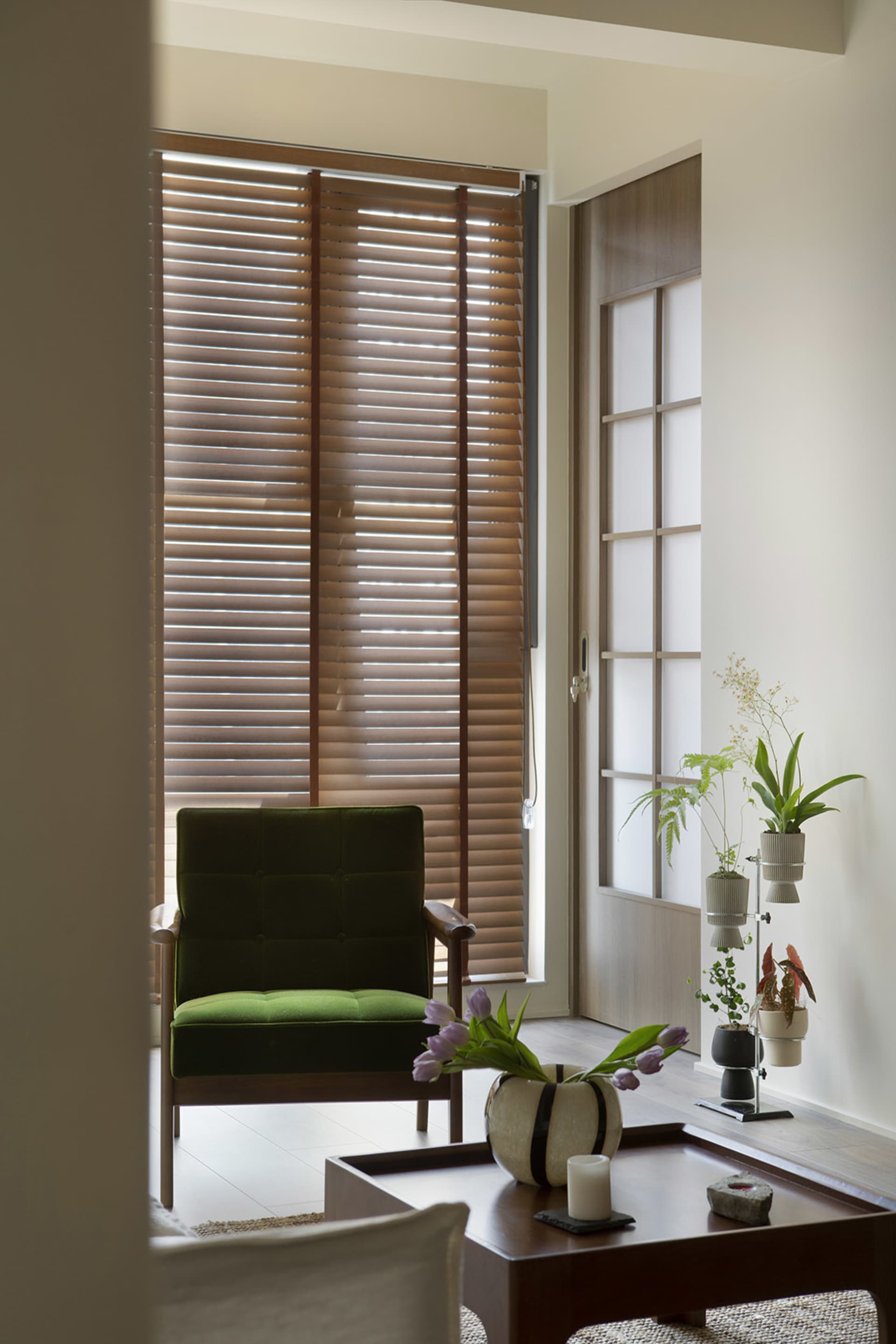 A minimalist living room with wooden blinds, a green accent chair, and a modern coffee table with plants and a vase of tulips