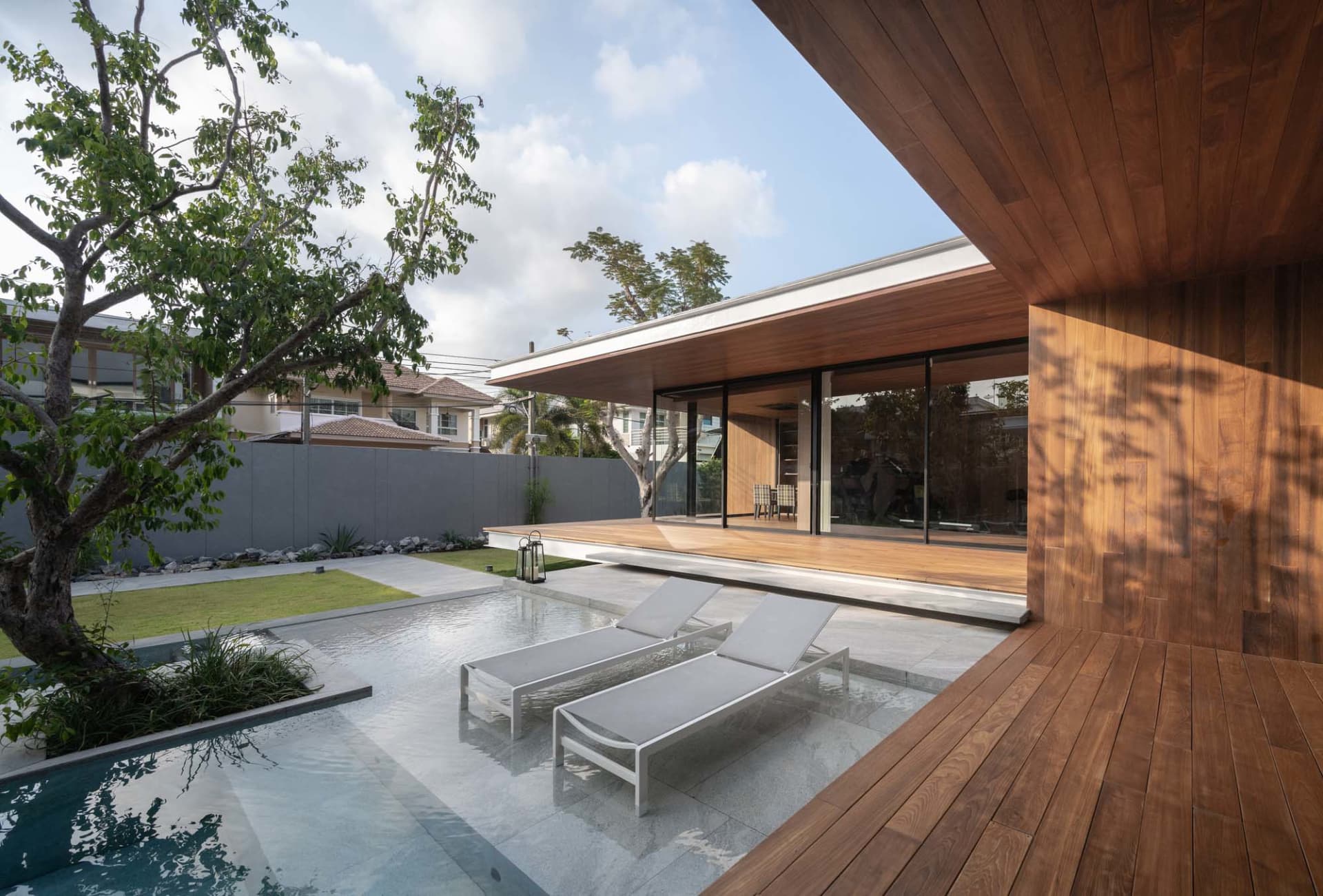 Modern residential home with wooden deck, glass walls, and concrete patio featuring outdoor furniture and a swimming pool sur