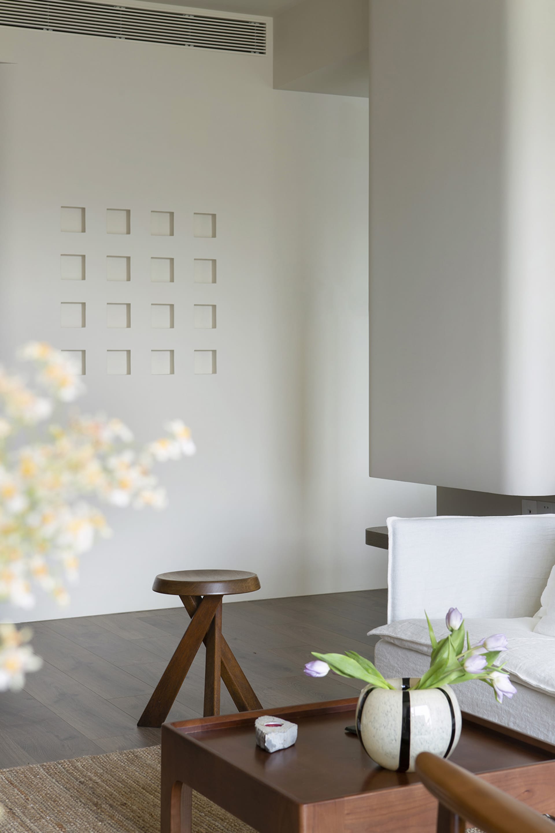 A modern minimalist living room with neutral tones, featuring a white sofa, wooden stool, and decorative vase with tulips.
