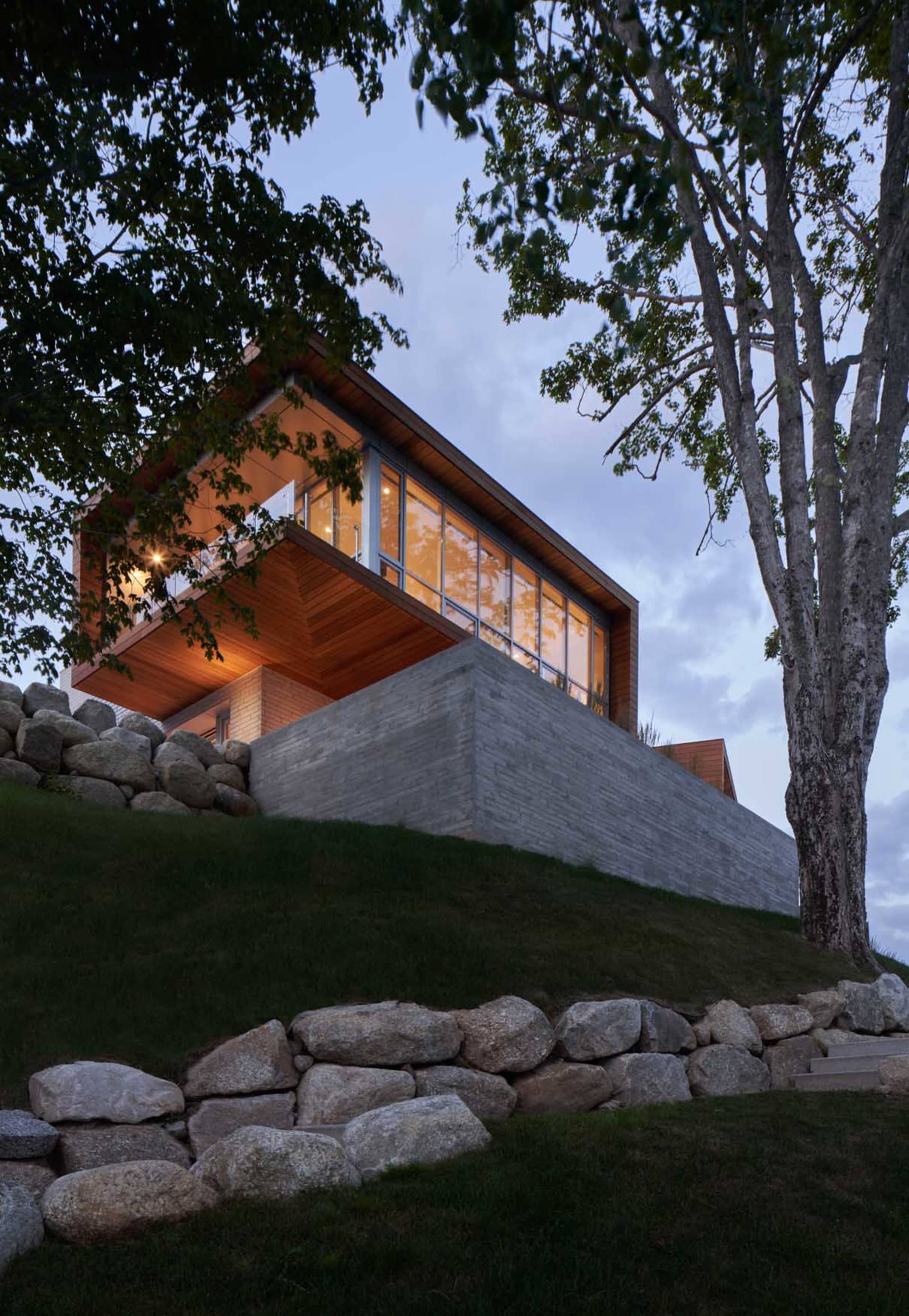 A modern house with a concrete exterior and large windows, surrounded by rocky ground and trees at dusk.
