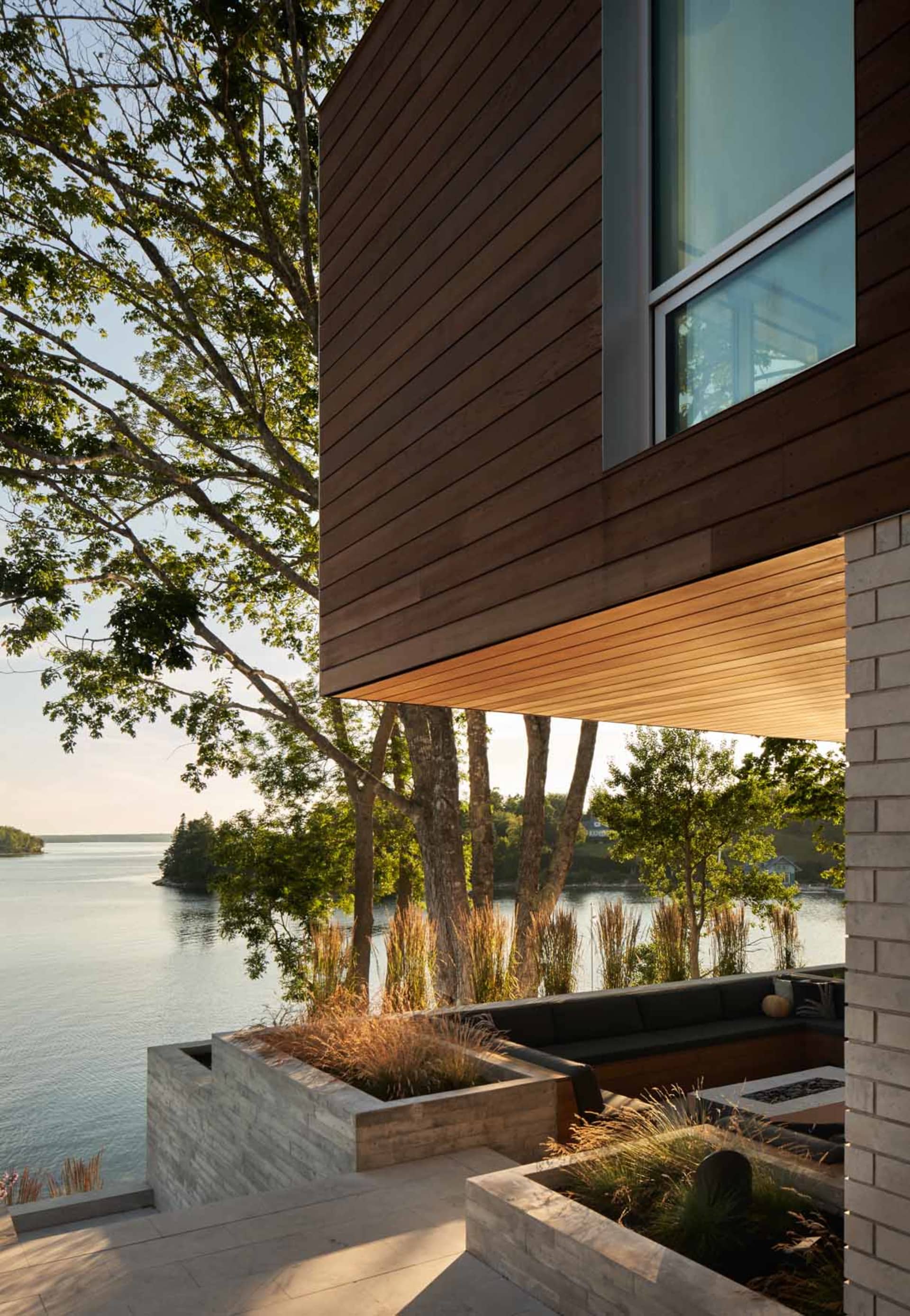 Modern residential exterior featuring wood siding, stone accents, and large glass windows overlooking water and greenery.