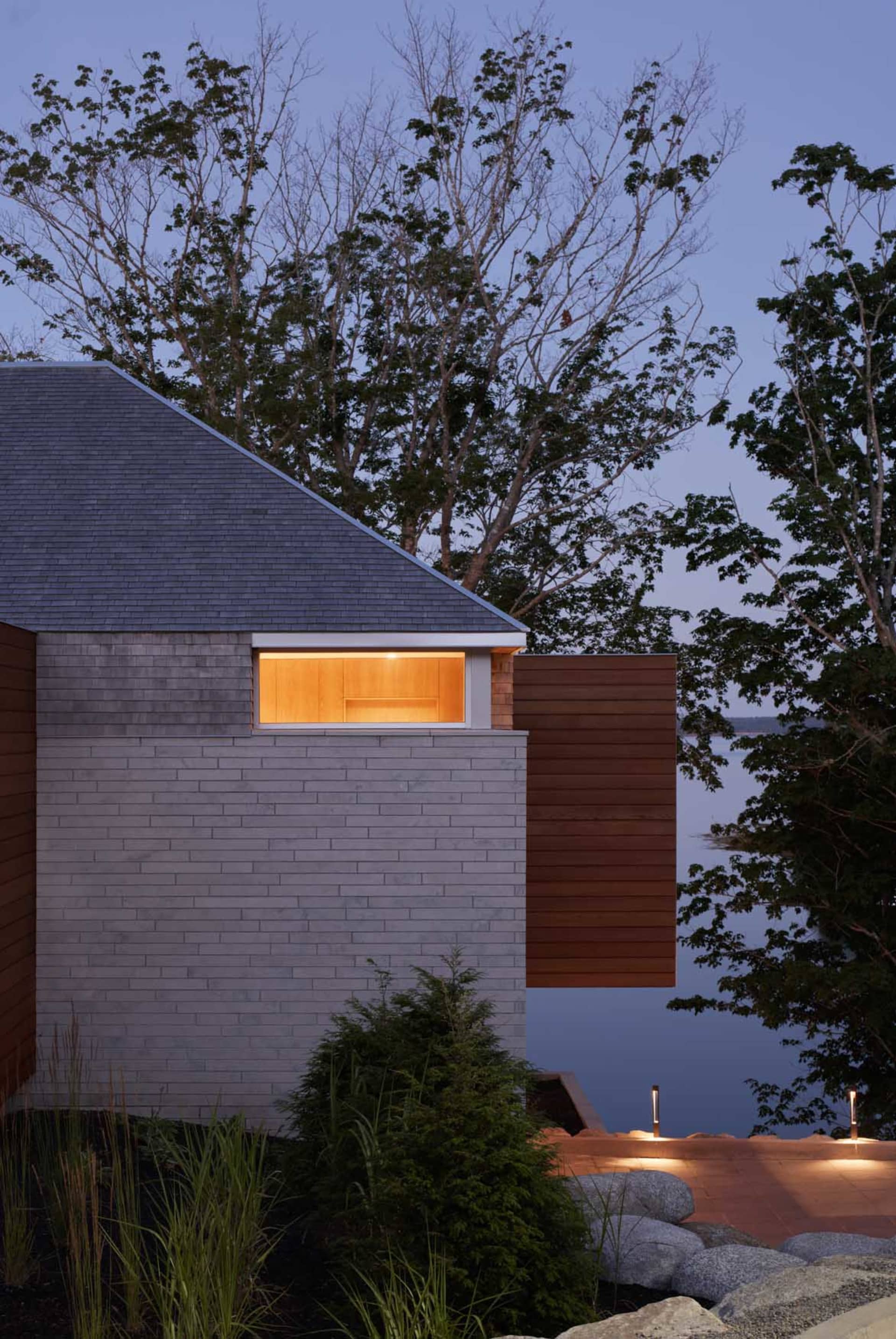 A modern residential home with a concrete exterior and wooden accents, featuring a warm indoor light at dusk.