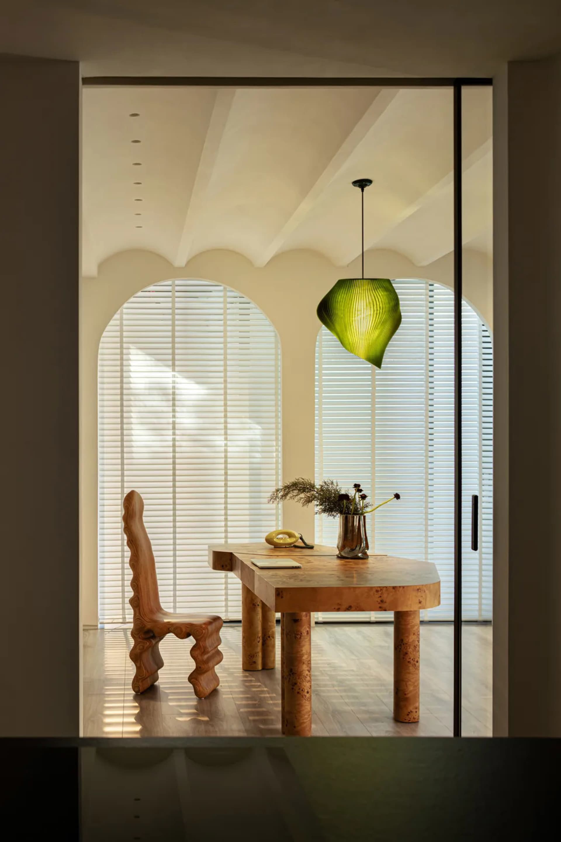 A modern home office featuring a wooden desk and chair, with a green pendant light and large windows.