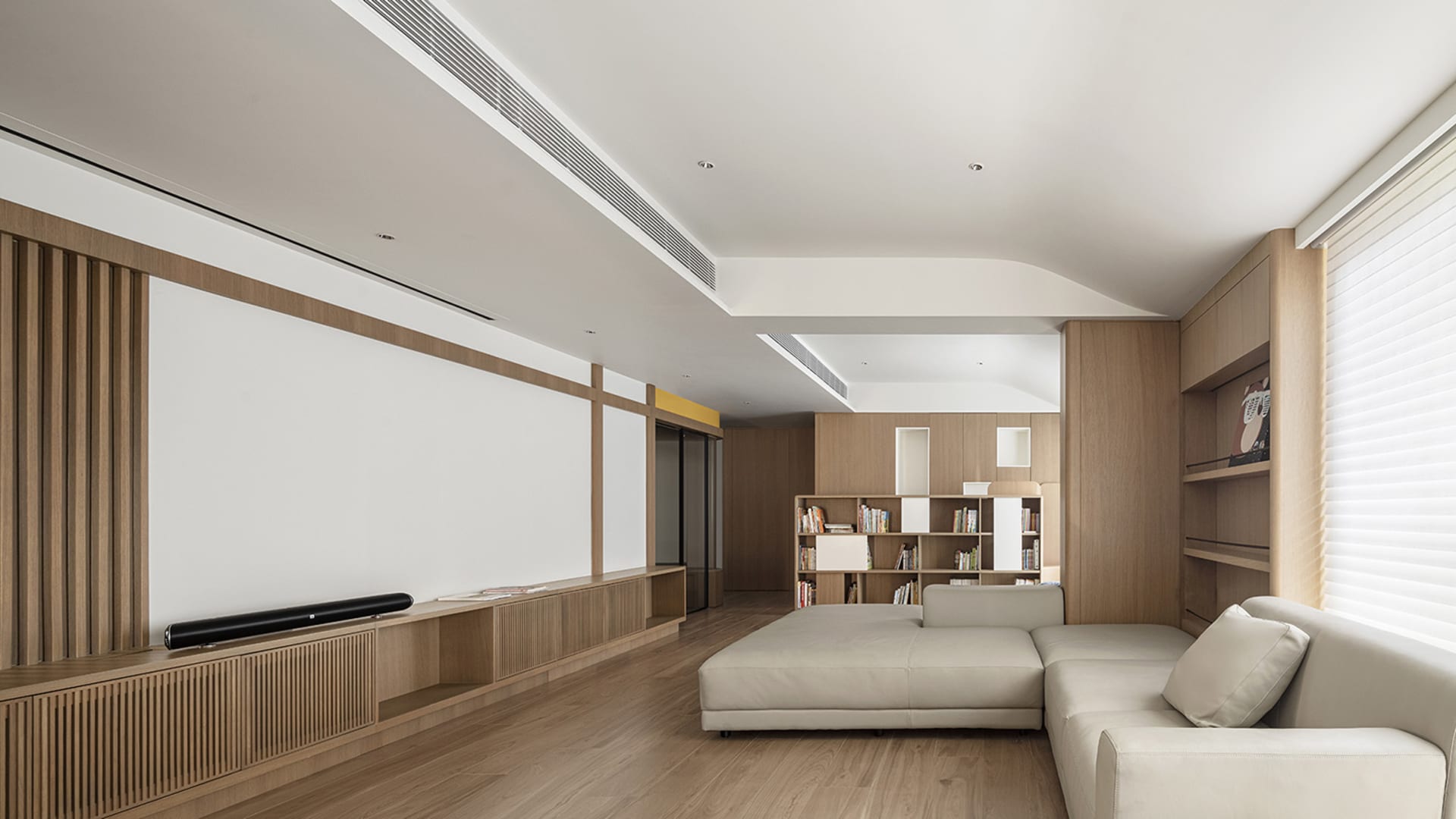 A spacious modern living room with wooden furniture, a white sofa, and a sleek floor-to-ceiling bookshelf.