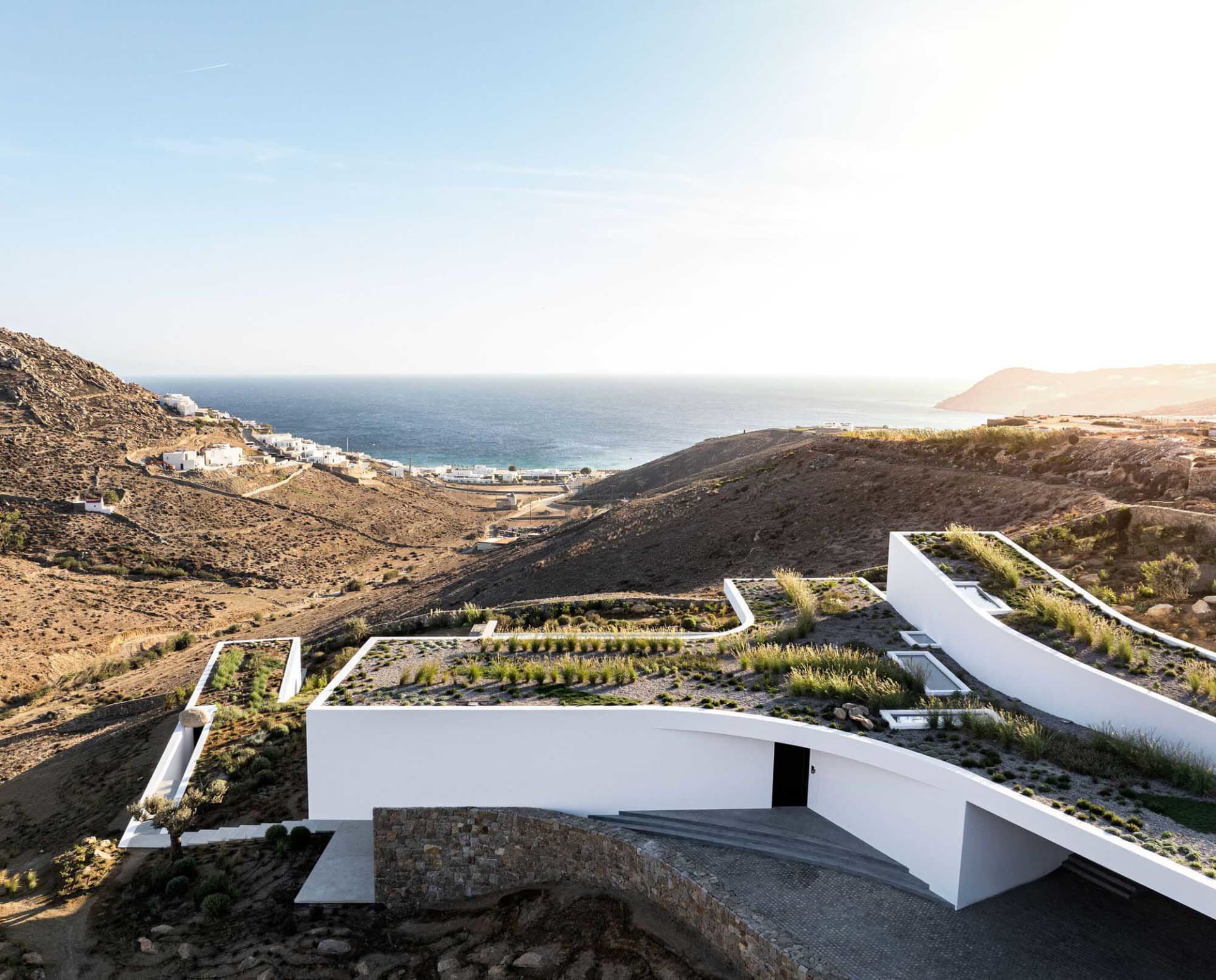 A modern, concrete residential building nestled in a rugged coastal landscape, featuring a rooftop garden overlooking the sea