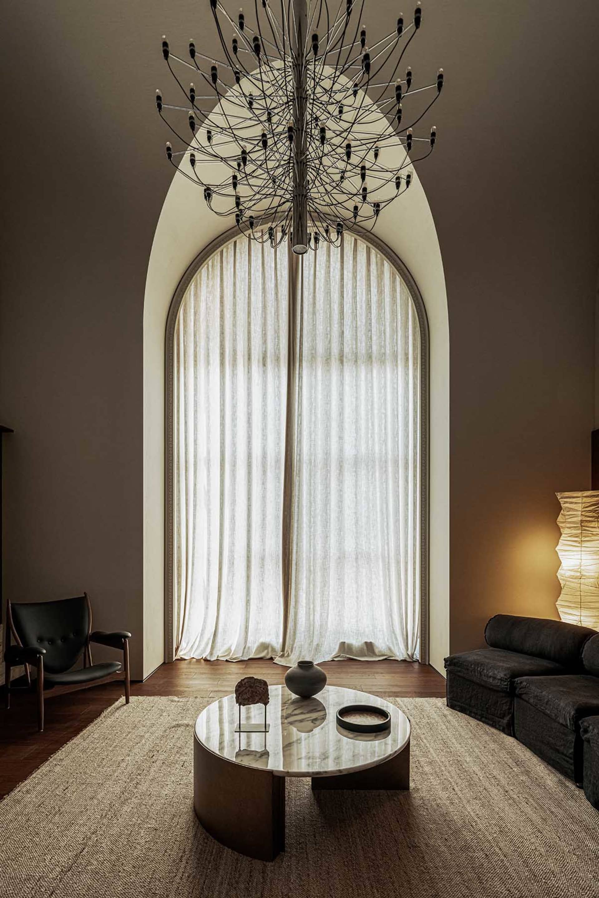 A modern living room featuring neutral-toned, organic materials and a large, arched window with white curtains.