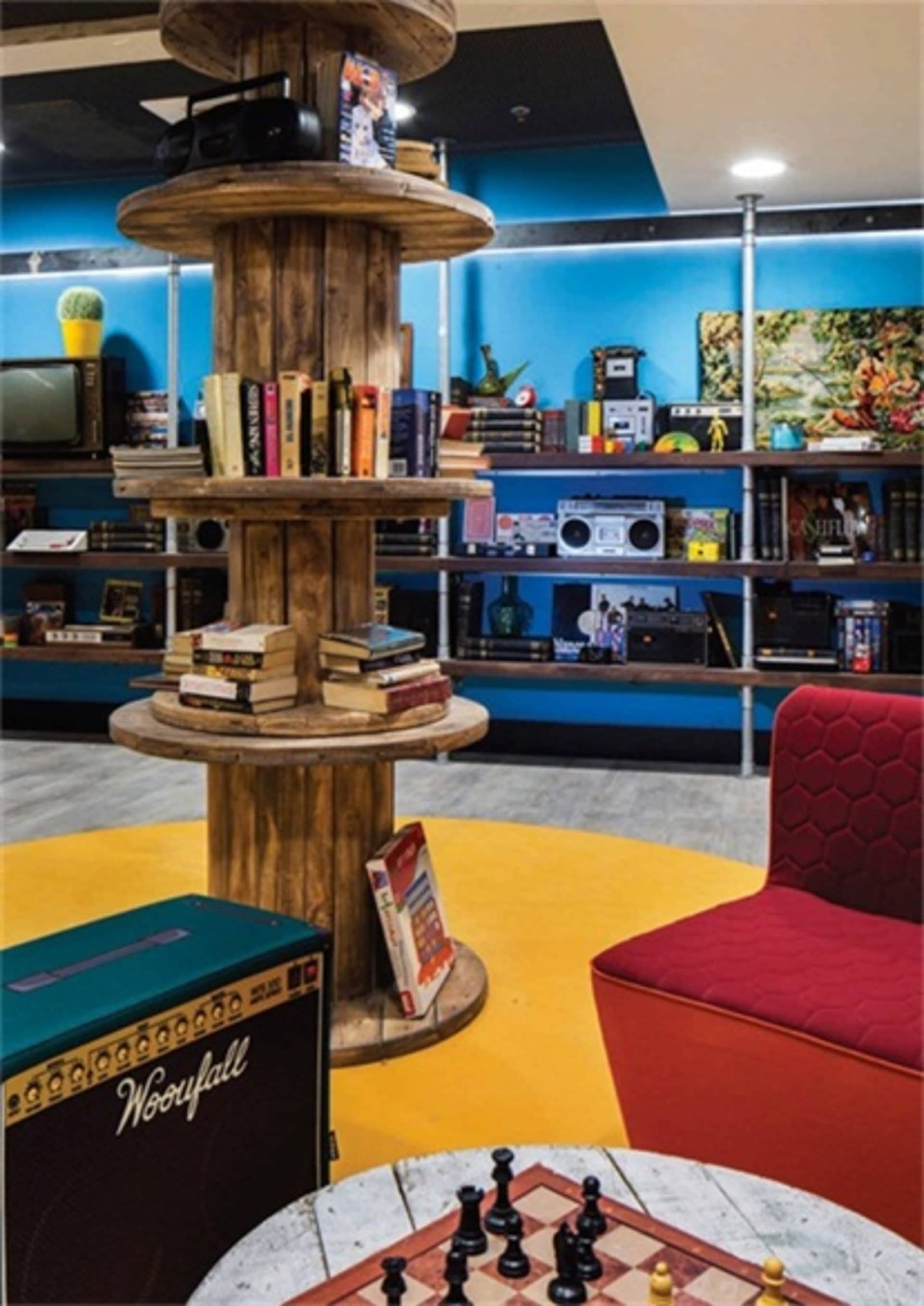 A cozy, industrial-style living space with a wooden bookshelf, red armchair, and chessboard.