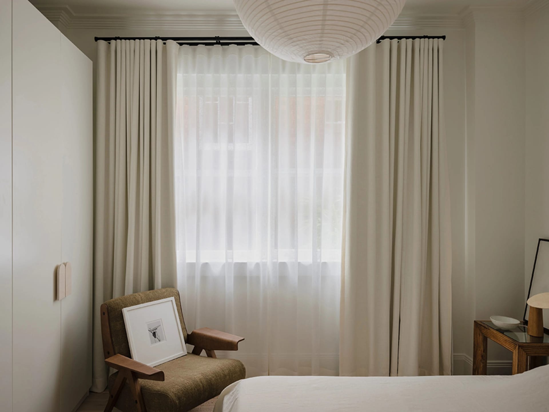 A modern, minimalist bedroom featuring a bed with white bedding, a wooden chair with a wall-mounted light fixture, and sheer