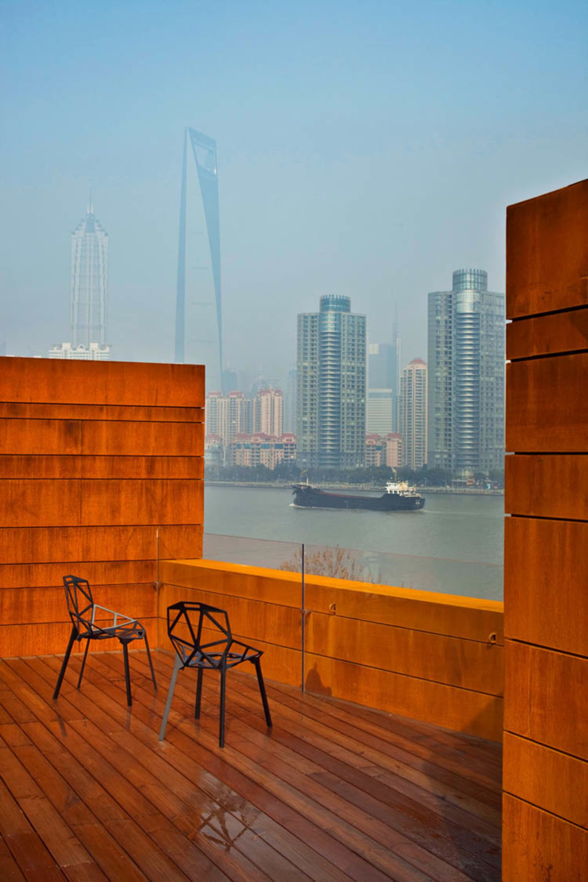 A wooden terrace overlooking modern urban buildings with metal accents and a ship on the water.