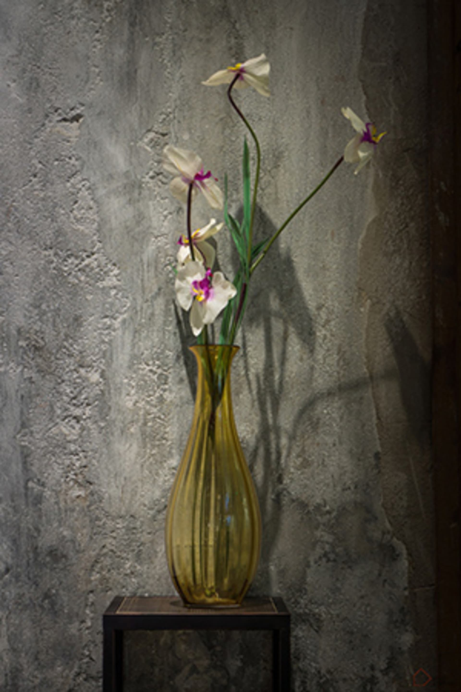 An industrial-style vase filled with flowers on a black table against a textured wall background.