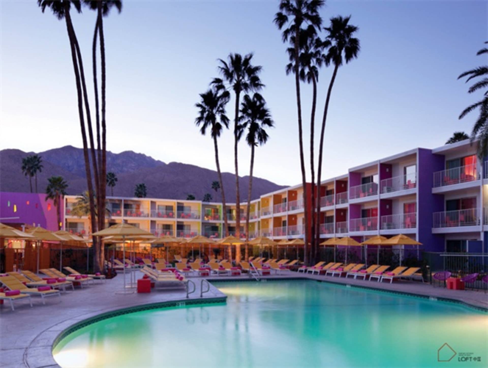 A resort scene featuring a large outdoor pool surrounded by palm trees, lounge chairs, and umbrellas with a mountain range in