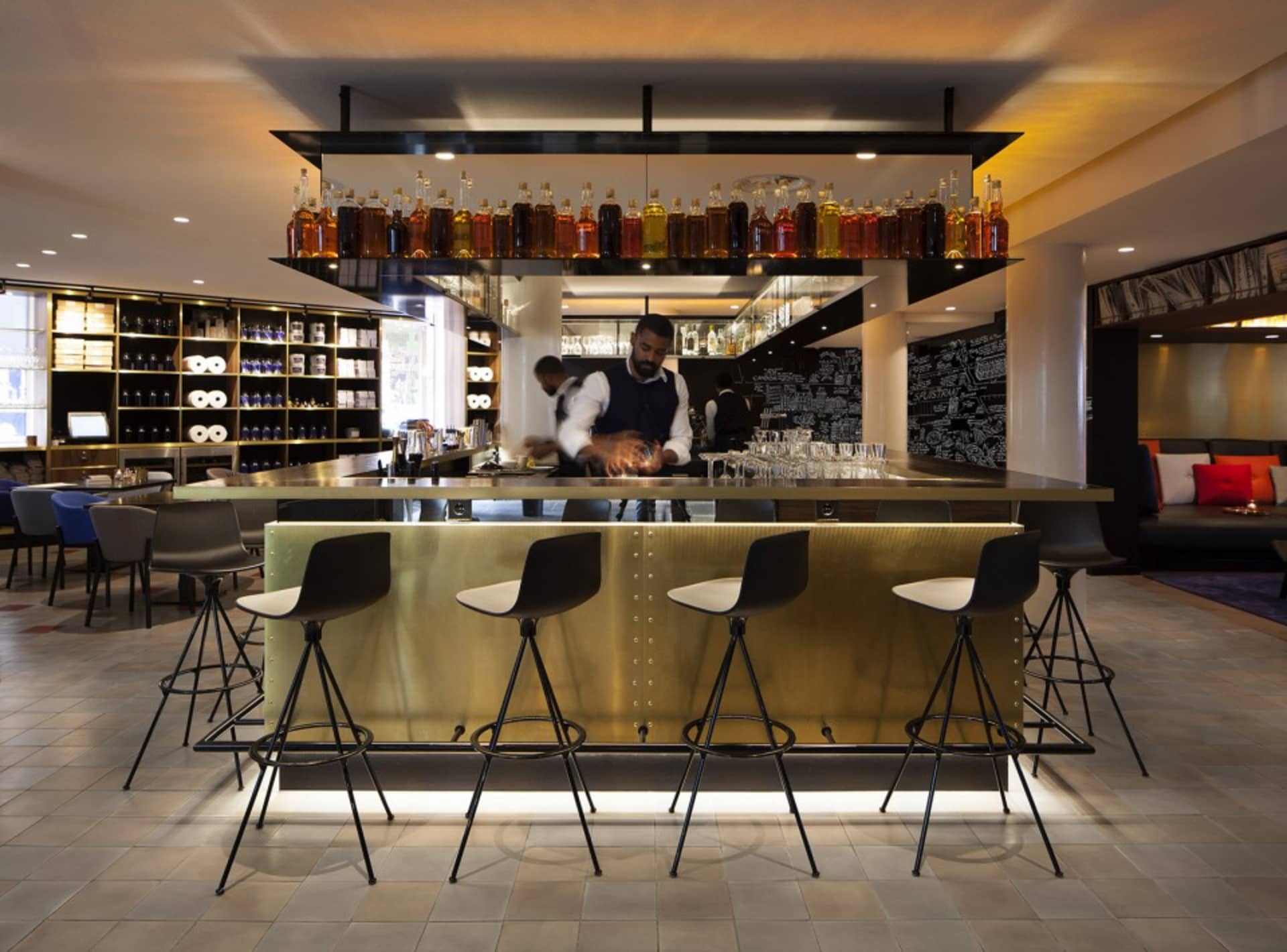 A stylish industrial hotel lobby featuring metallic bar stools, a brass counter, and a variety of alcohol bottles lined up on