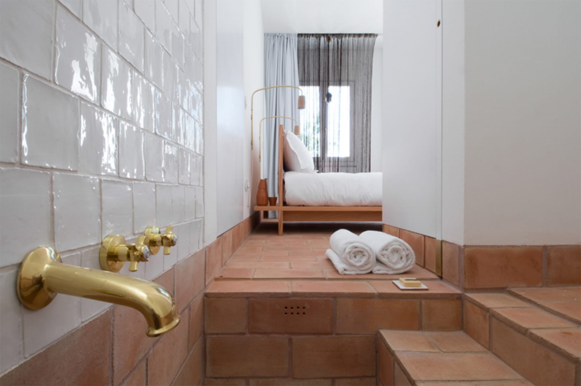 A minimalist bedroom featuring white tiled walls and terracotta tiles on the floor, with a bed and towels.