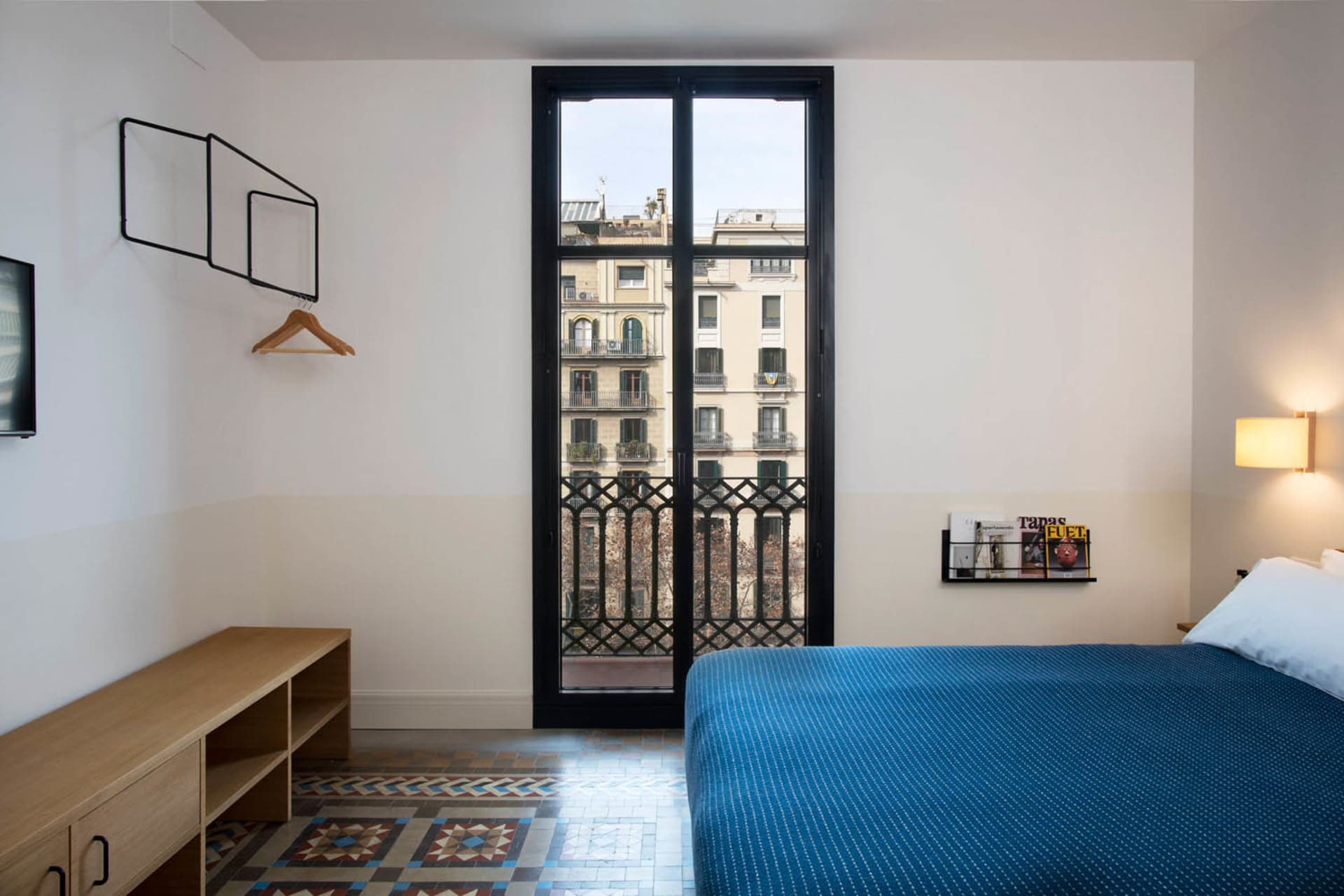 A minimalist bedroom featuring a blue bed with a wooden headboard, white walls, and a large window with a metal railing.