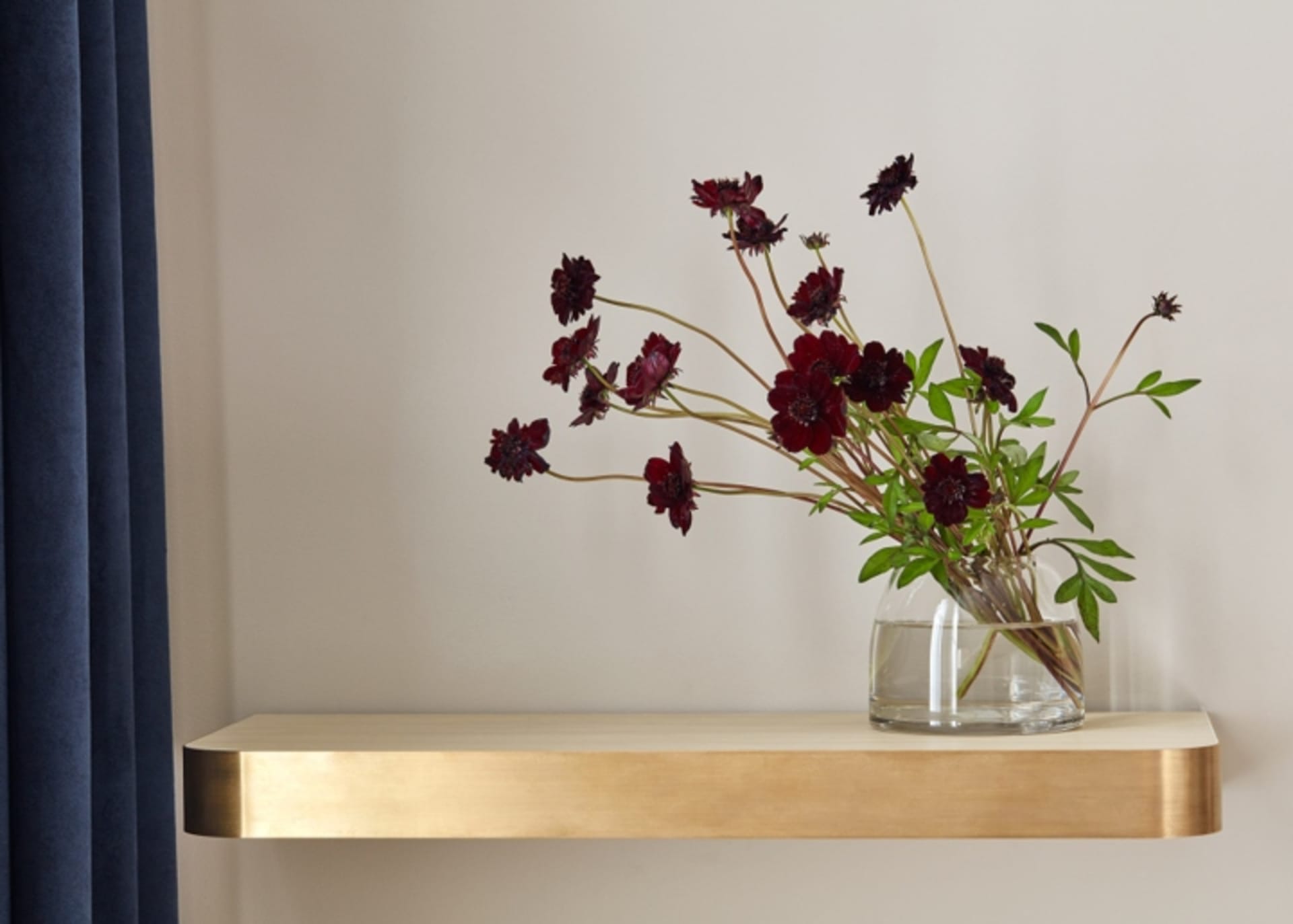 Glass vase with dark flowers on a brass shelf against a white wall.