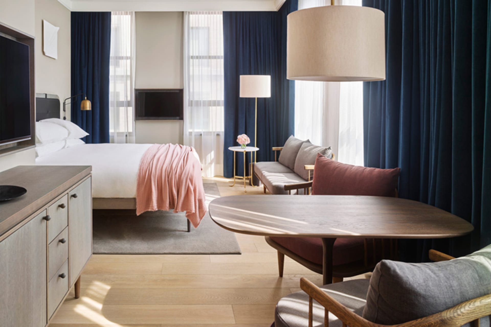 A cozy modern bedroom with a bed dressed in soft pink bedding, a wooden table, and a plush sofa adorned with cushions.