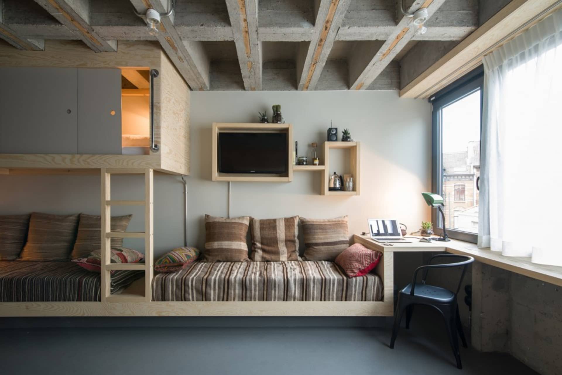 A modern living space featuring a concrete bunk bed, a built-in sofa, a small desk, and a window with a view of the city.