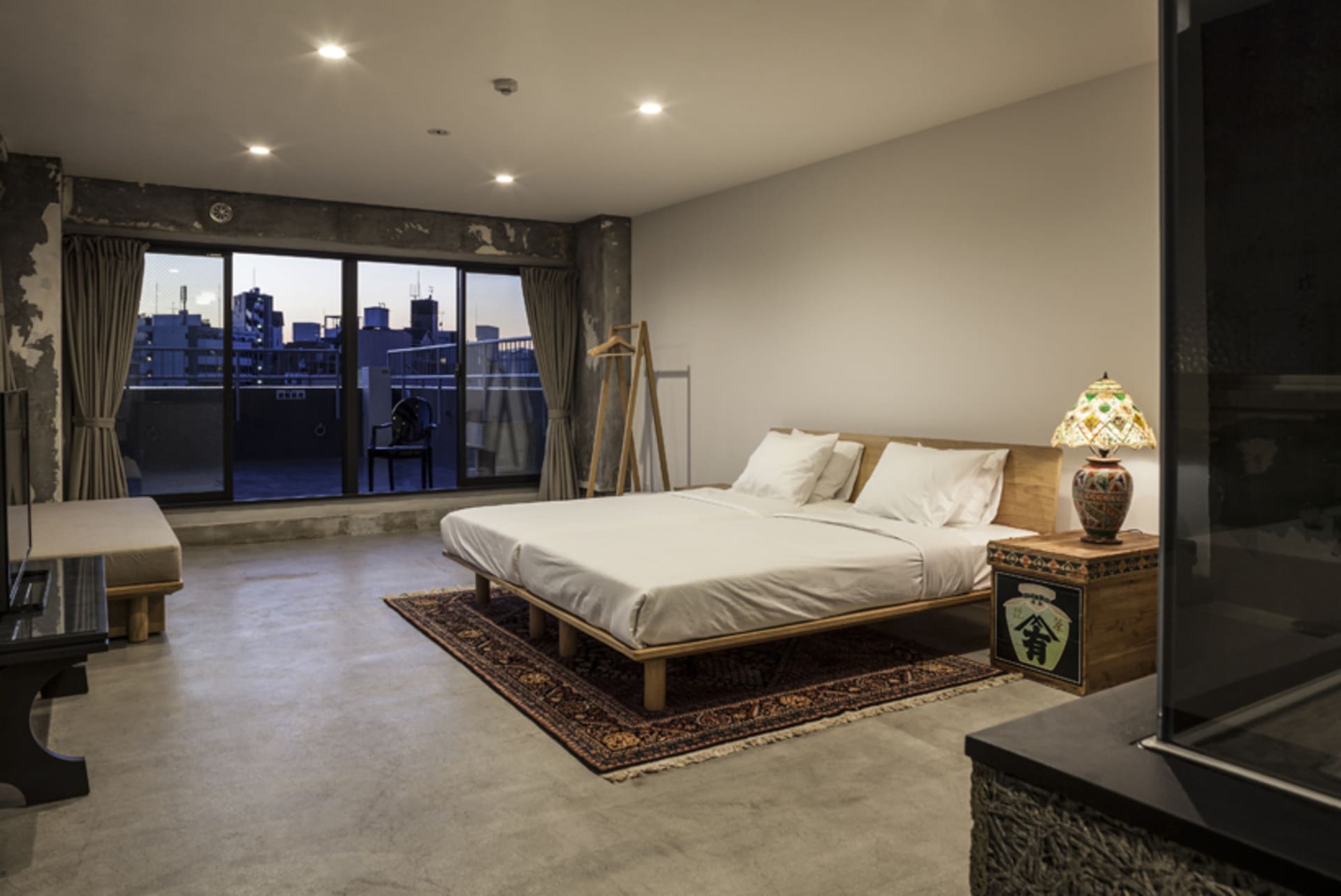 Modern bedroom with concrete floor, wooden bed frame, and rug, featuring a cityscape view from a window