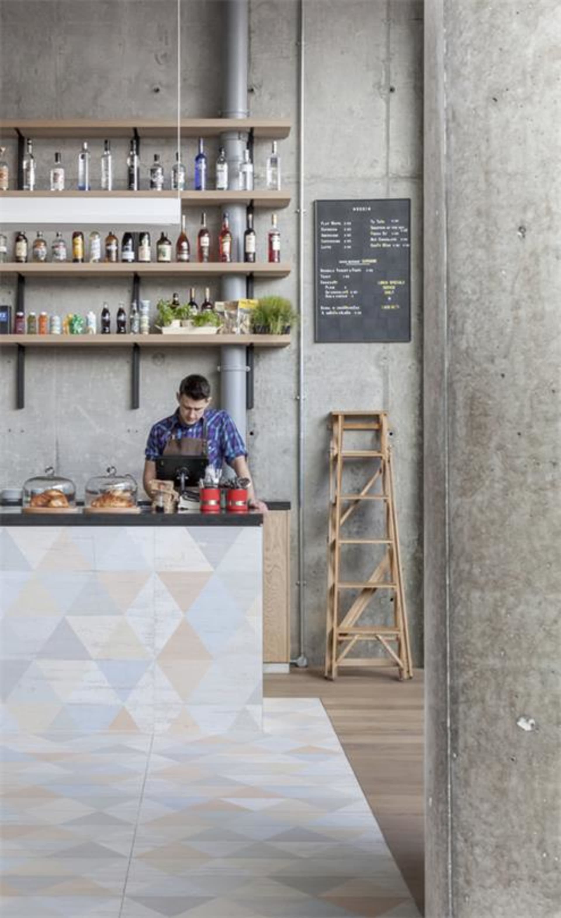 A worker stands behind a coffee bar adorned with various bottles and plants, with a menu chalkboard and a wooden step ladder