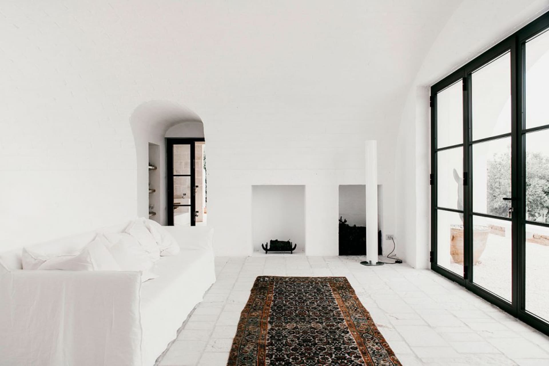 A minimalist living room featuring a concrete floor and walls, with a white sofa, a patterned rug, and a fireplace.